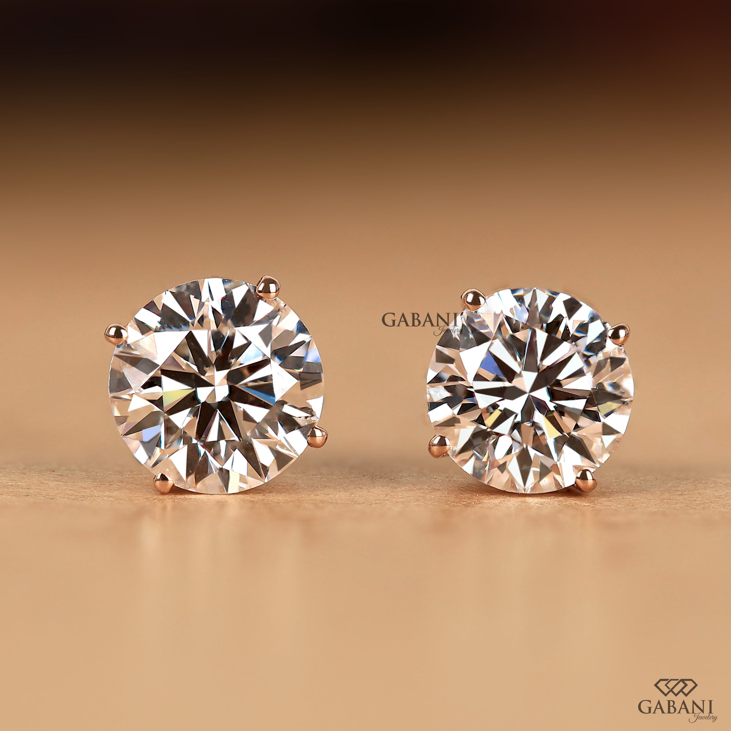 A pair of round-cut diamond stud earrings in a rose gold setting, displayed on a warm brown gradient background. The brand name 'GABANI' is visible as a watermark and logo in the lower right corner.