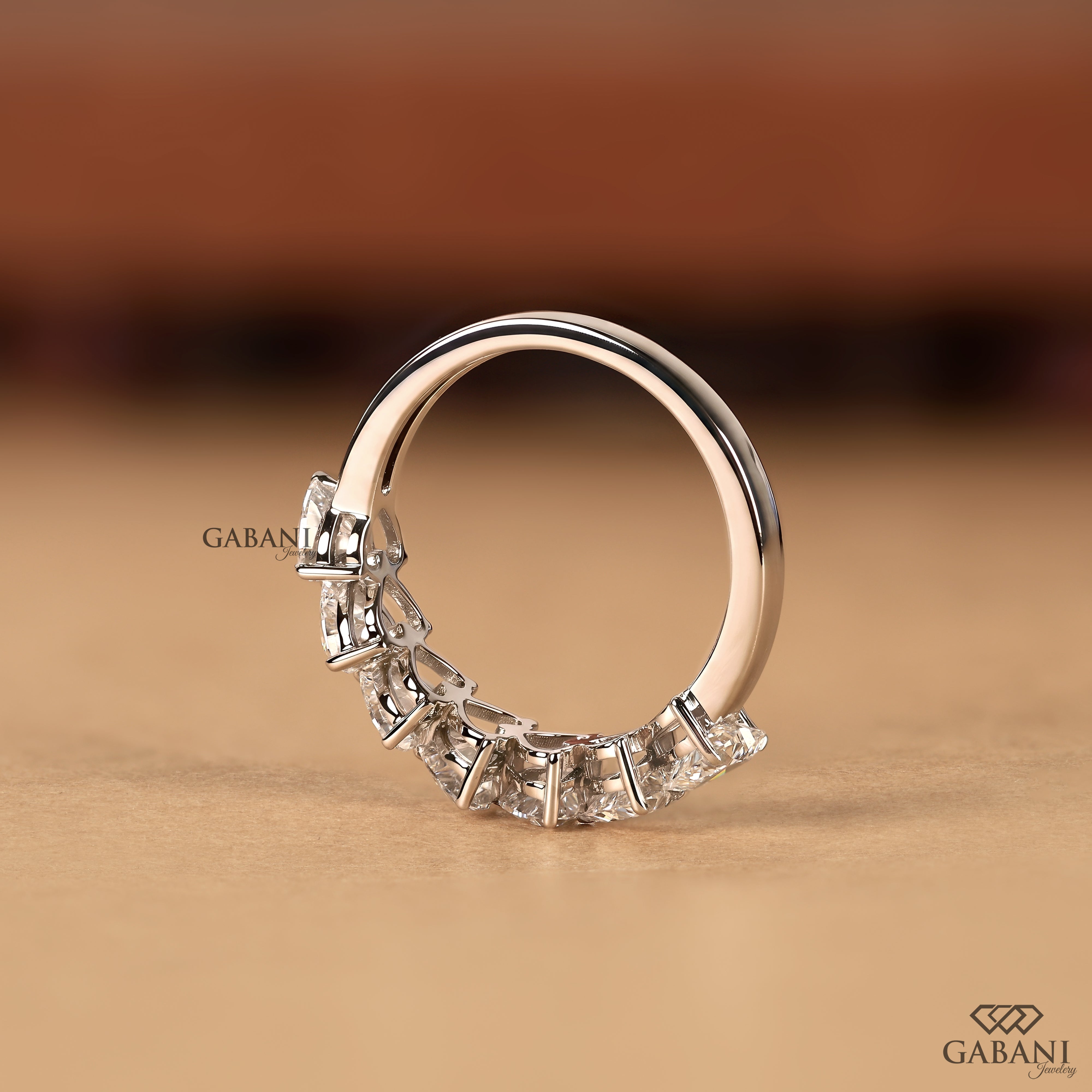 A silver-toned ring with a curved row of clear rectangular gemstones on the front, photographed on a warm beige surface with a blurred brown background. The brand name GABANI is visible as a watermark and logo.