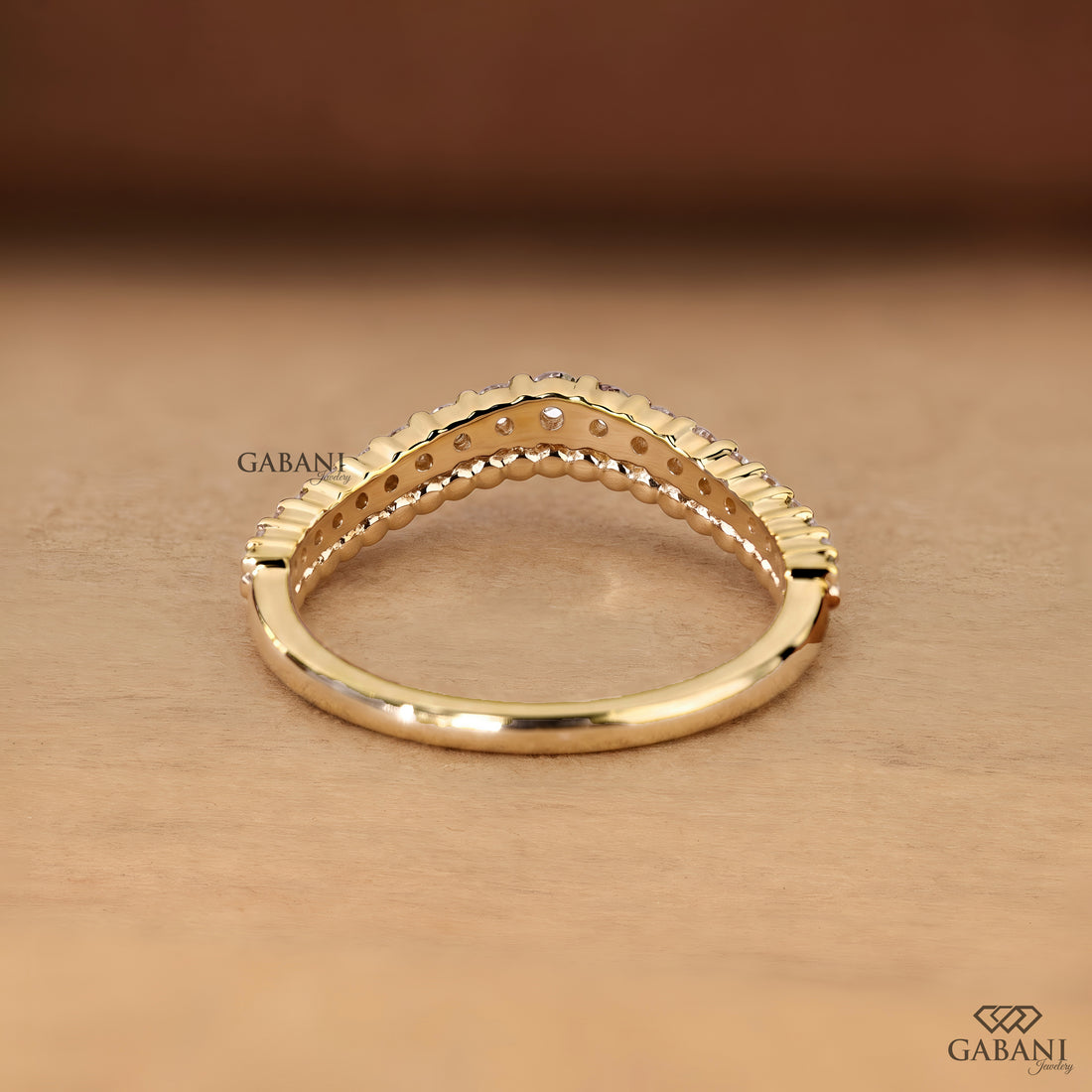 A gold ring with a textured beaded top band and a smooth shank, displayed on a warm brown wooden surface. The brand name 'GABANI' is visible as a watermark.