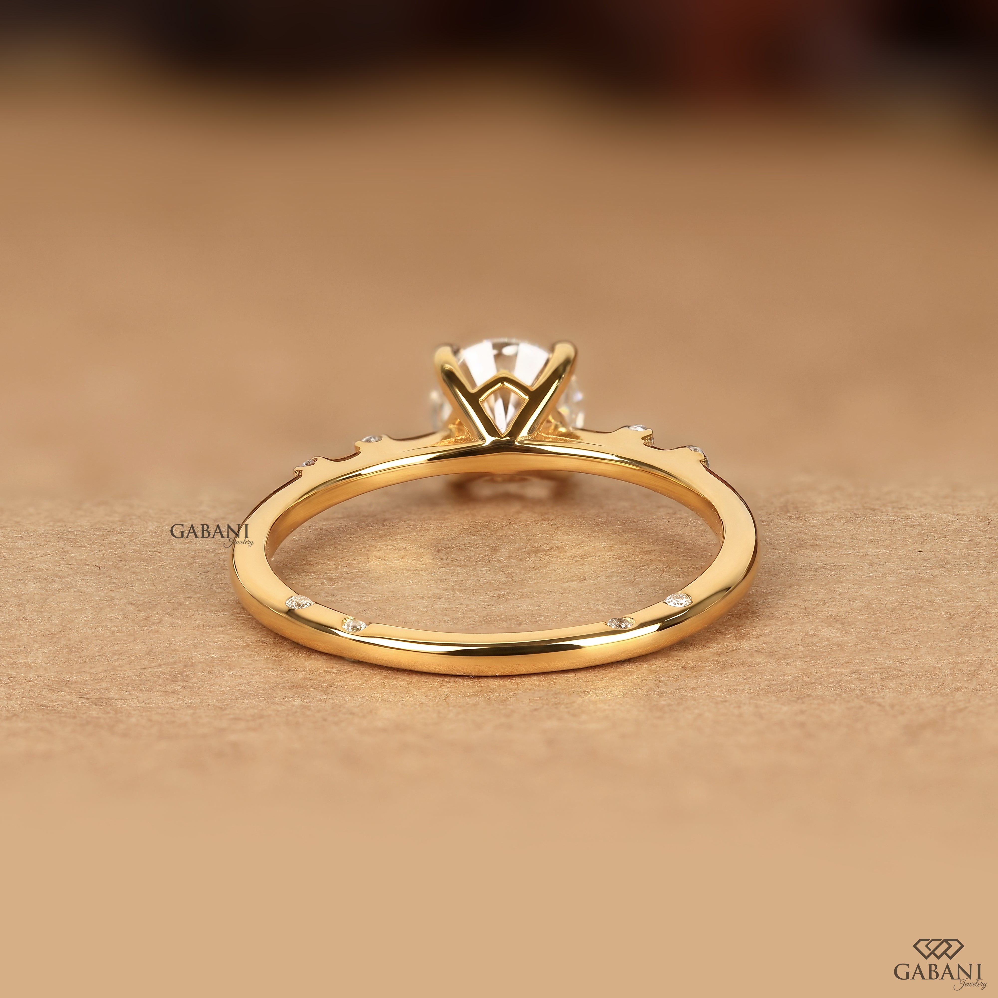 A gold ring with a multi-prong setting and small accent stones, shown on a warm beige textured background. The brand name GABANI is visible as a watermark.