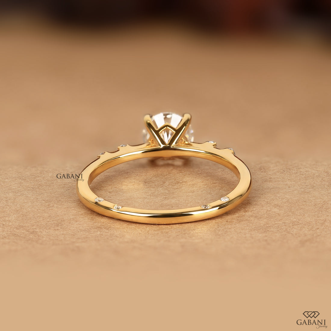 A gold ring with a multi-prong setting and small accent stones, shown on a warm beige textured background. The brand name GABANI is visible as a watermark.