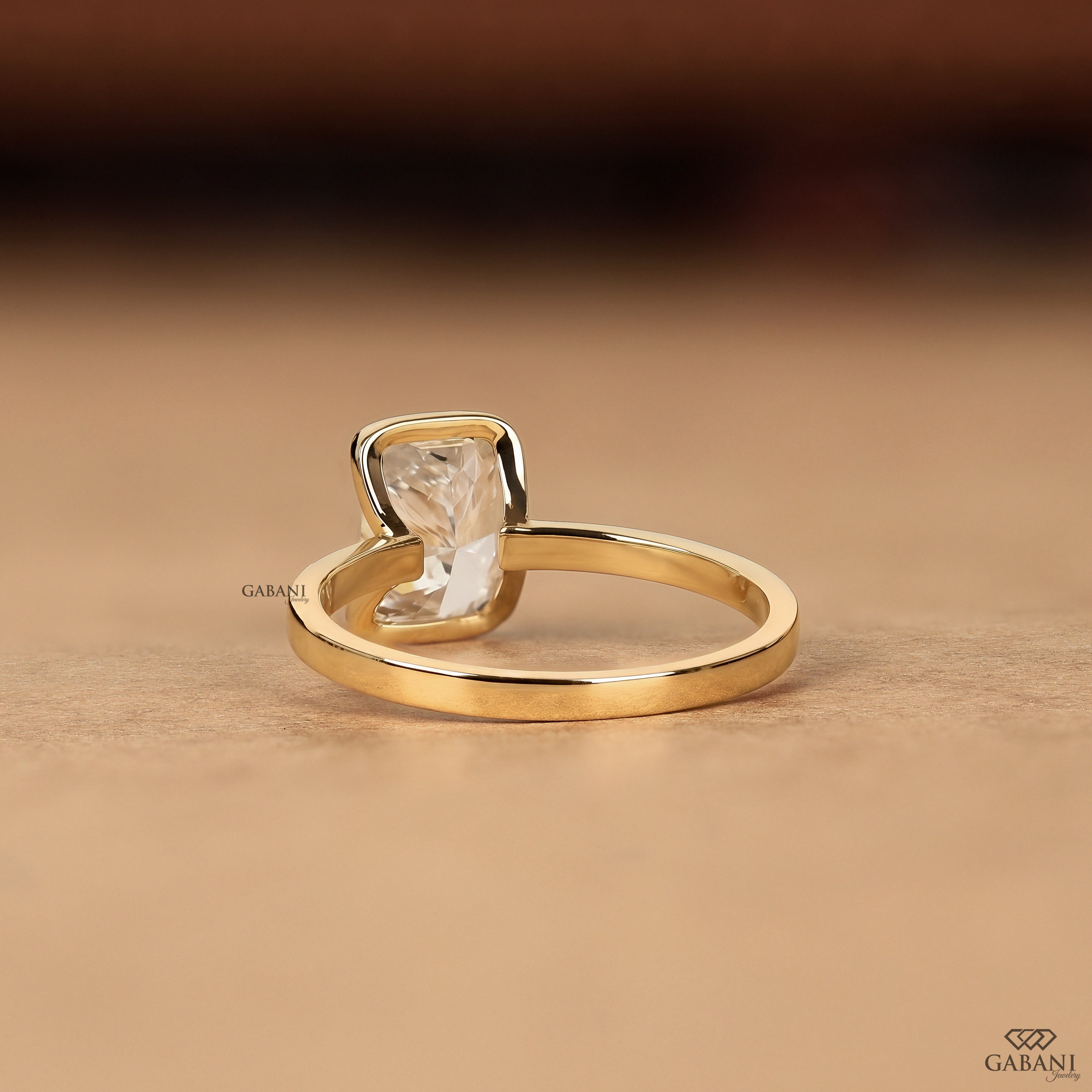A gold ring with a rectangular clear gemstone in a bezel setting, resting on a textured beige surface. The brand name 'GABANI' is visible as a watermark.