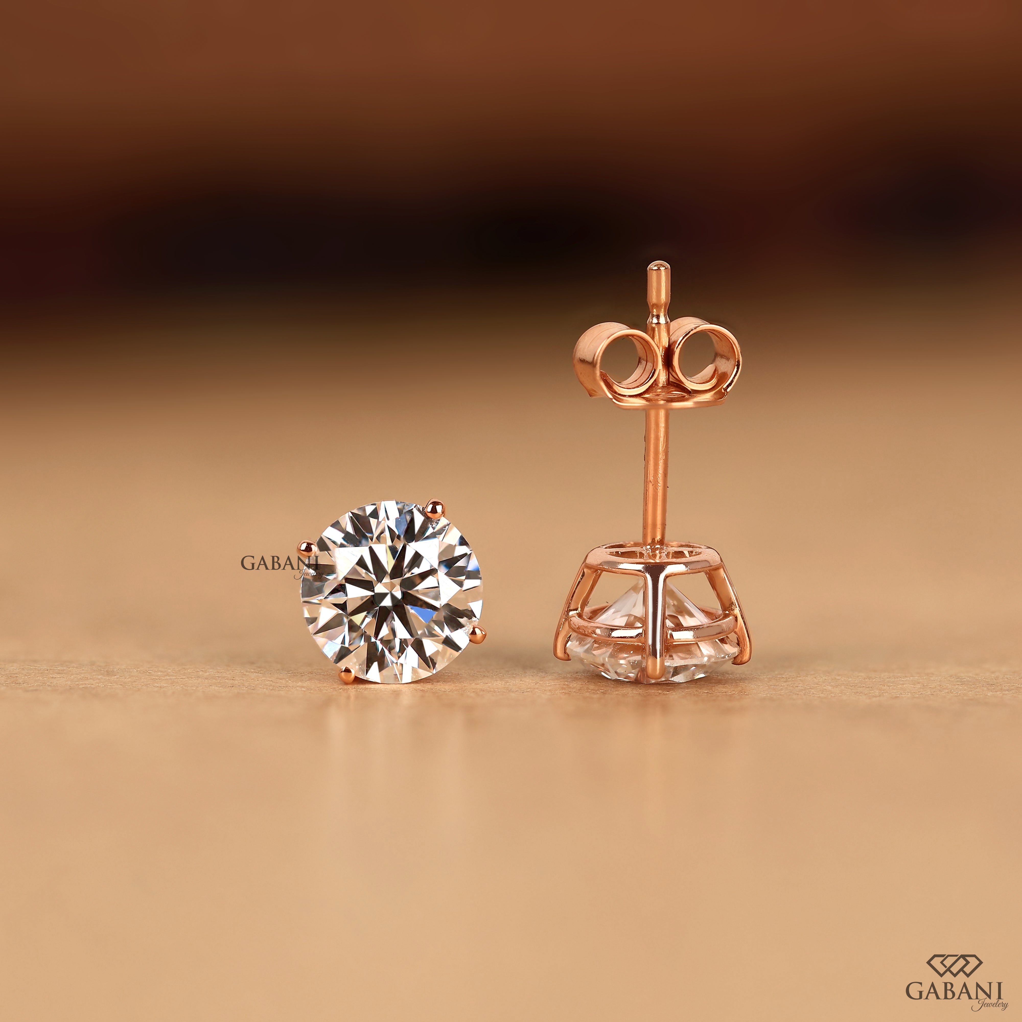 A pair of rose gold stud earrings, with one earring shown front-facing to display a round brilliant-cut clear gemstone and the other shown from the side/back to display the rose gold post and butterfly clasp. The brand name 'GABANI' is visible as a watermark.