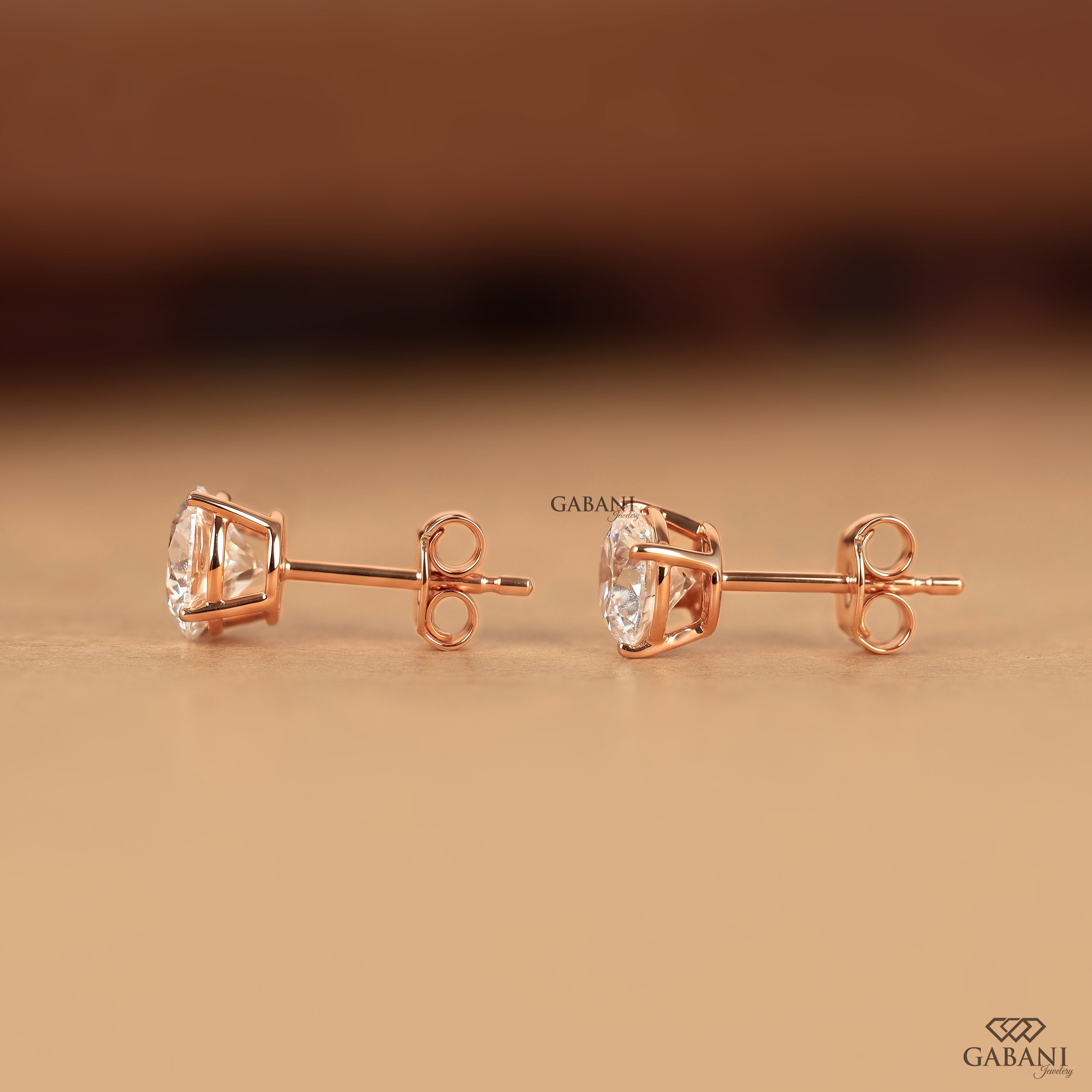 A pair of rose gold stud earrings with clear gemstones, displayed on a warm beige surface with a blurred brown background. The brand name 'GABANI' is visible as a watermark and in the bottom right corner.