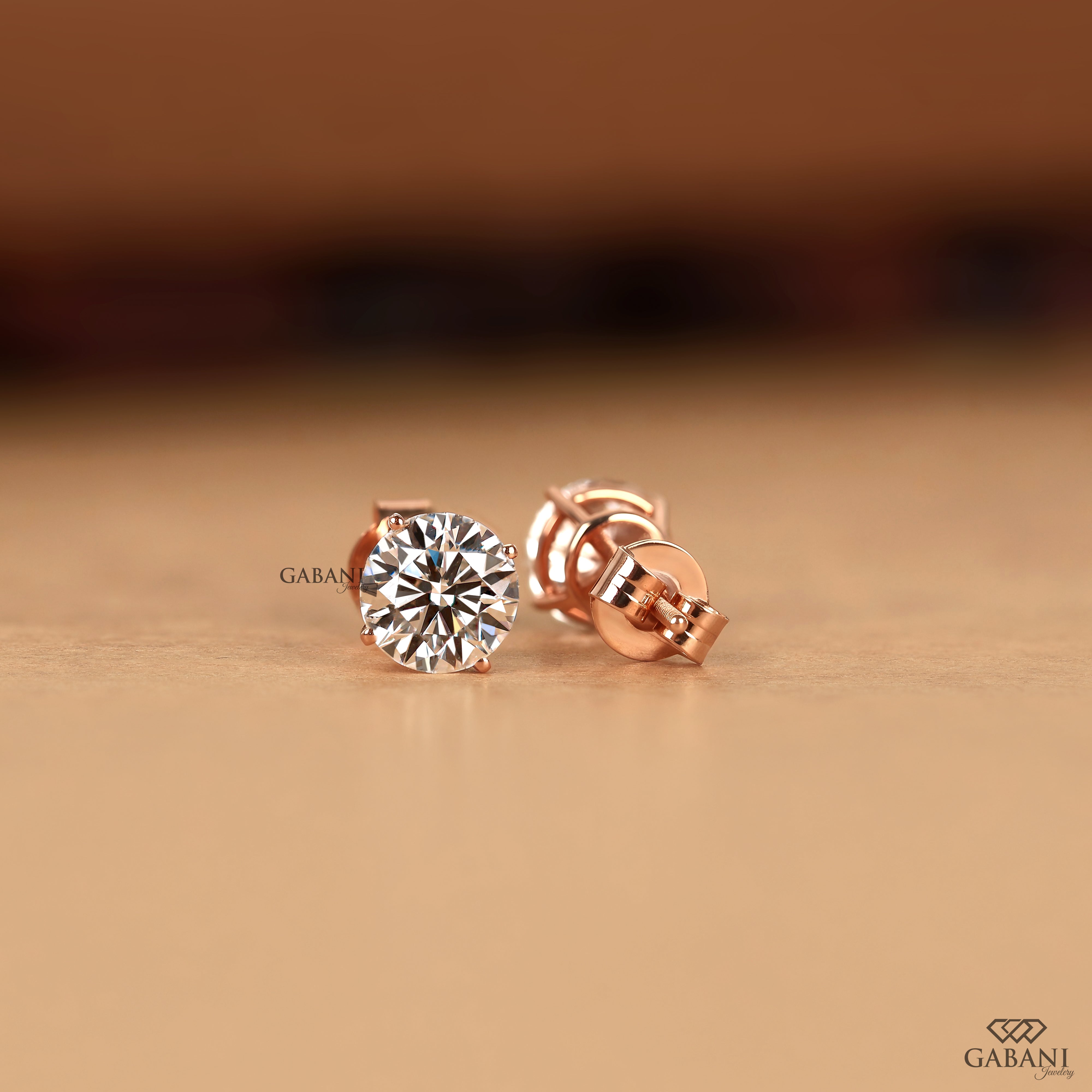 A pair of rose gold stud earrings with round-cut clear gemstones, displayed on a warm beige surface with a blurred brown background. The brand name 'GABANI' is visible as a watermark.