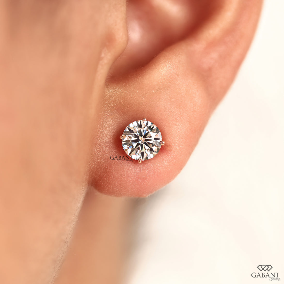 A close-up of a person's ear wearing a single round brilliant-cut diamond stud earring in a silver-toned setting. The brand name GABANI is visible near the earring and in the bottom right corner.