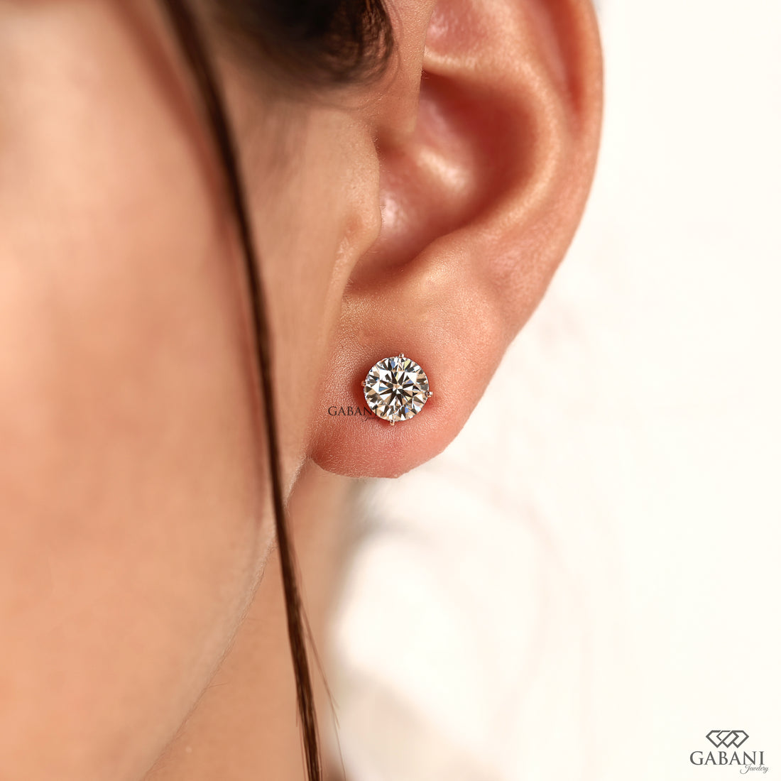 A close-up of a person's ear wearing a round brilliant-cut diamond stud earring in a silver setting, with the brand name GABANI visible near the earring and in the bottom right corner.