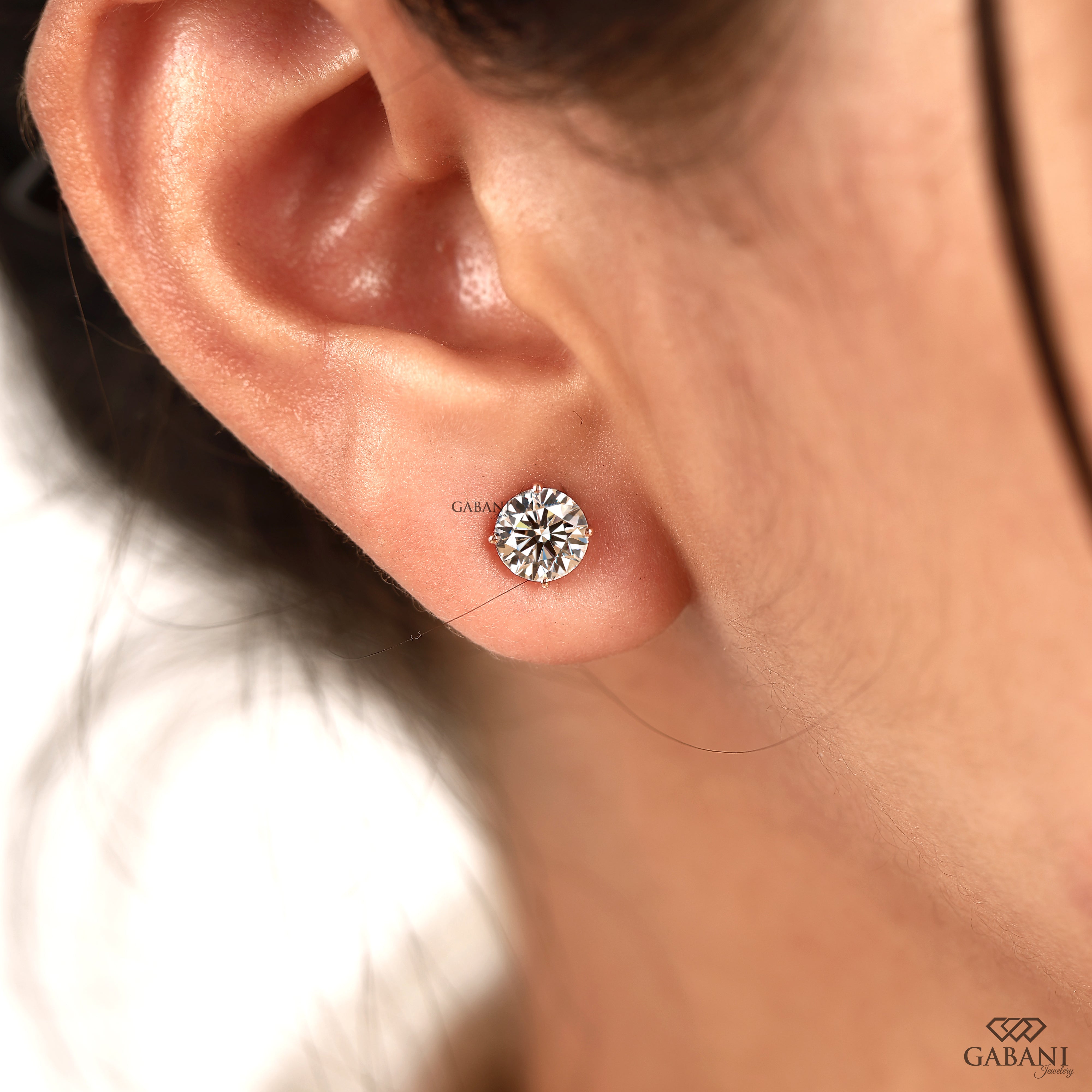 A close-up of a person's ear wearing a small round brilliant-cut diamond stud earring in a silver setting. The brand name 'GABANI' is visible as a watermark in the bottom right corner and faintly near the earring.