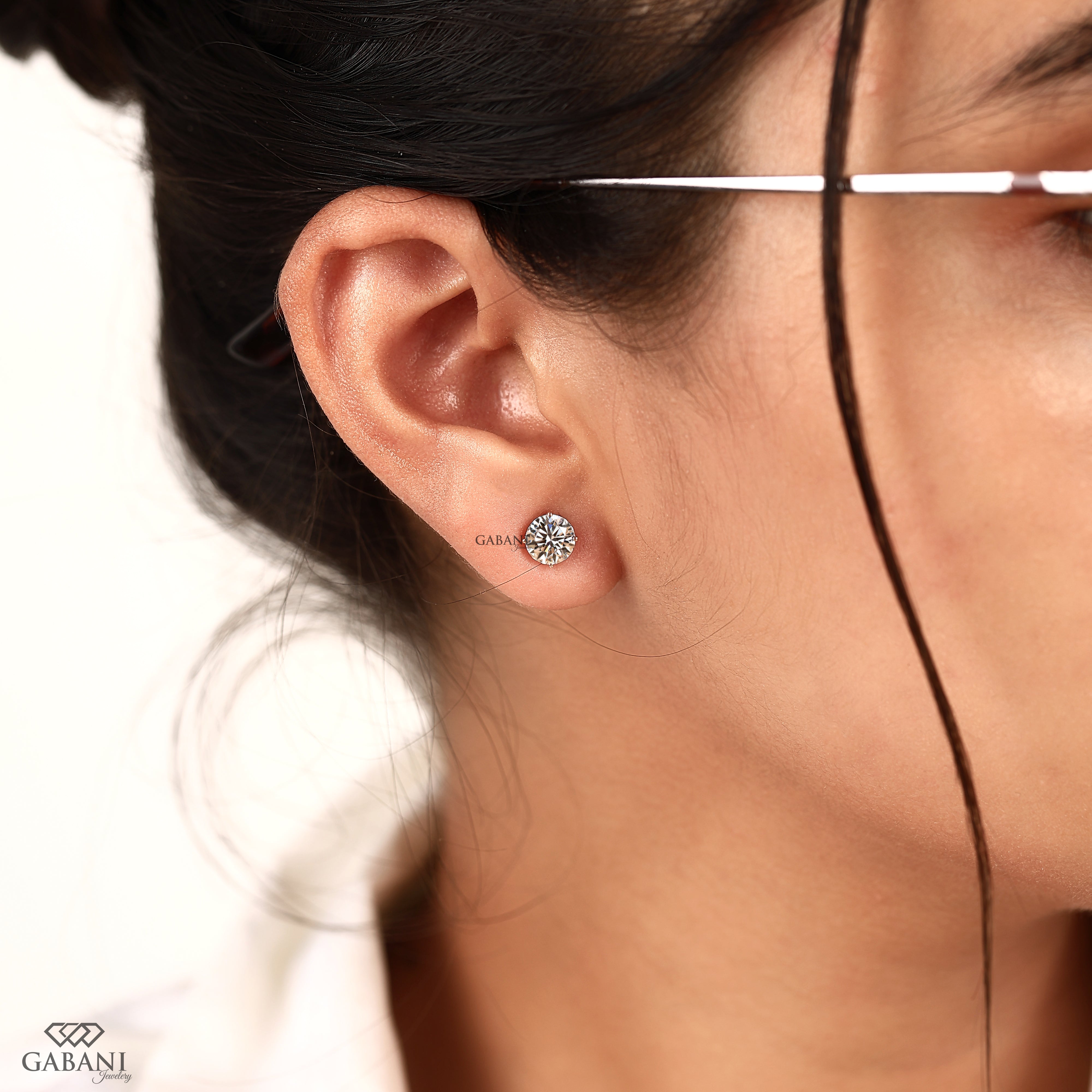 A close-up of a woman's ear wearing a small round stud earring with a sparkling clear gemstone. The brand name 'GABANI' is printed near the earring and appears again in the bottom-left corner.