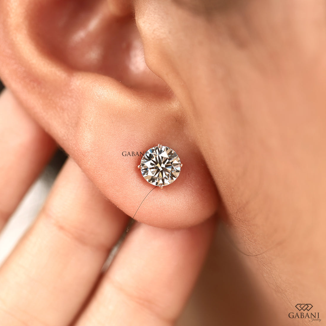 A close-up of a round brilliant-cut diamond stud earring in a silver-toned setting, worn on a person's ear lobe. The brand name 'GABANI' is printed near the earring, and the 'GABANI' logo is visible in the bottom right corner.