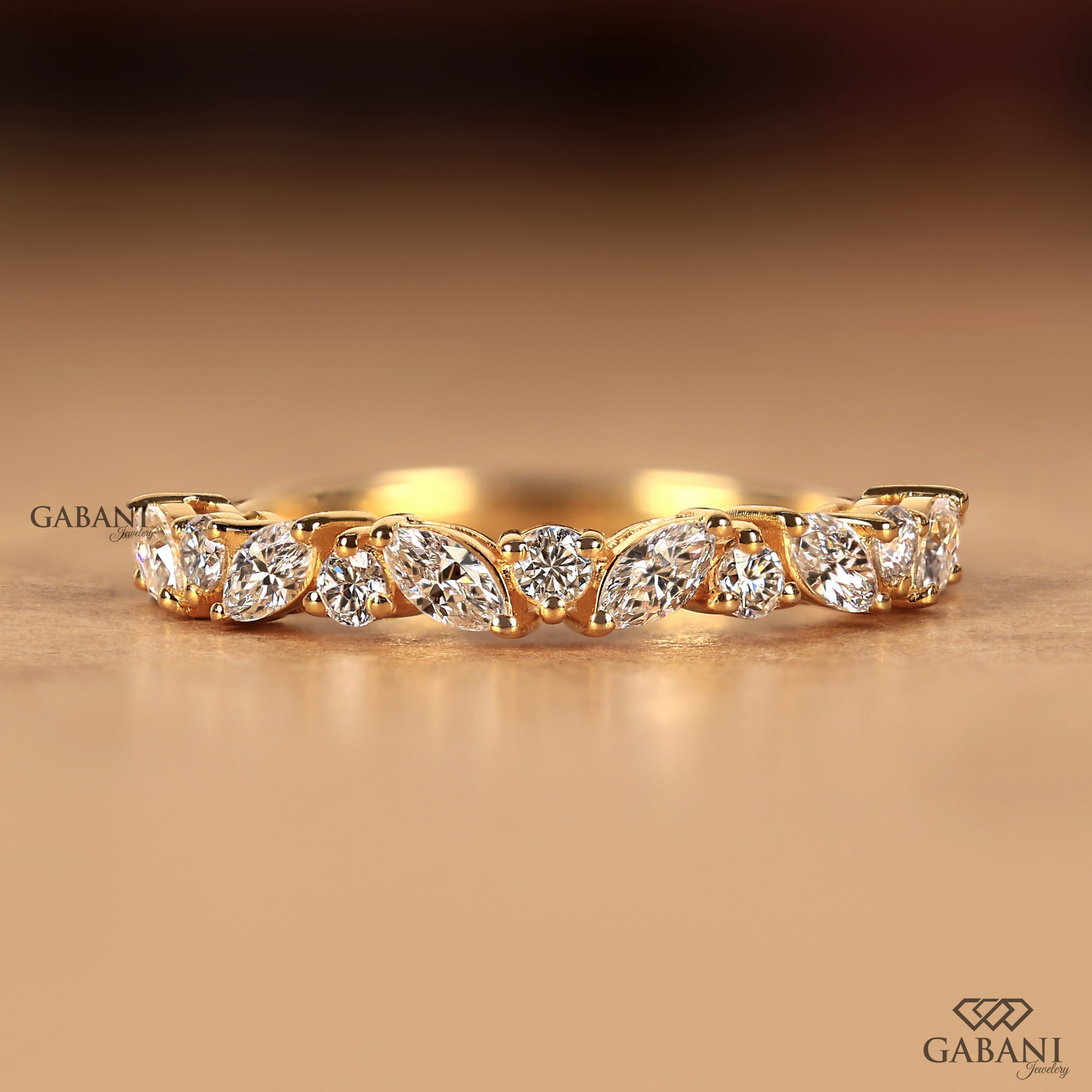 A yellow gold eternity ring featuring a row of marquise-cut and round brilliant-cut diamonds, displayed on a warm beige surface with a soft-focus background. The brand name 'GABANI' is visible as a watermark on the left and a logo in the bottom right corner.