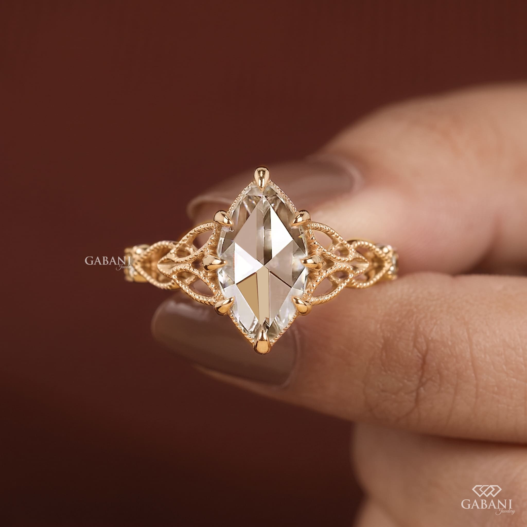 A gold ring with a marquise-cut clear gemstone and an intricate filigree band, held between fingers against a dark brown background.