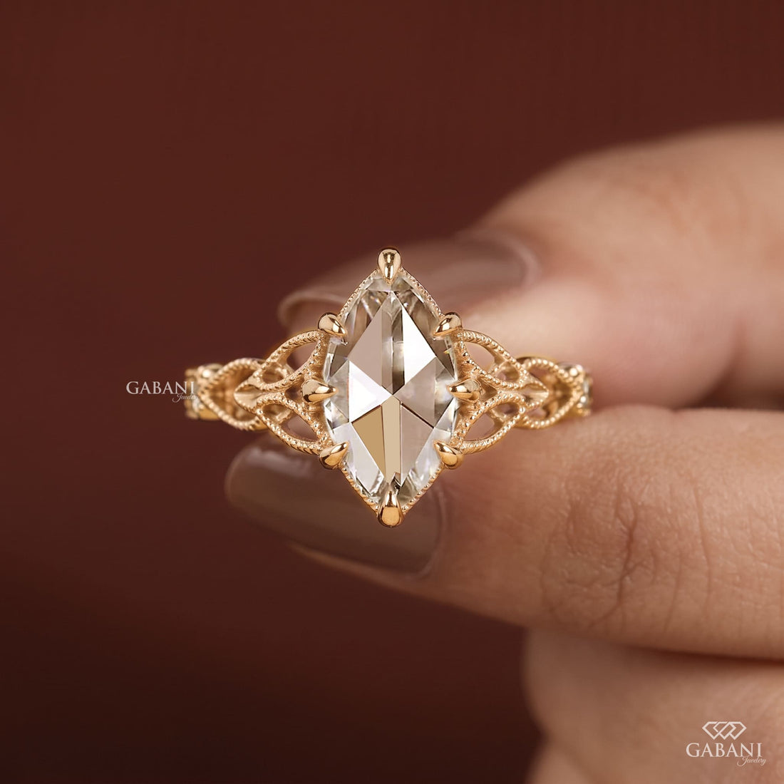 A gold ring with a marquise-cut clear gemstone and an intricate filigree band, held between fingers against a dark brown background.