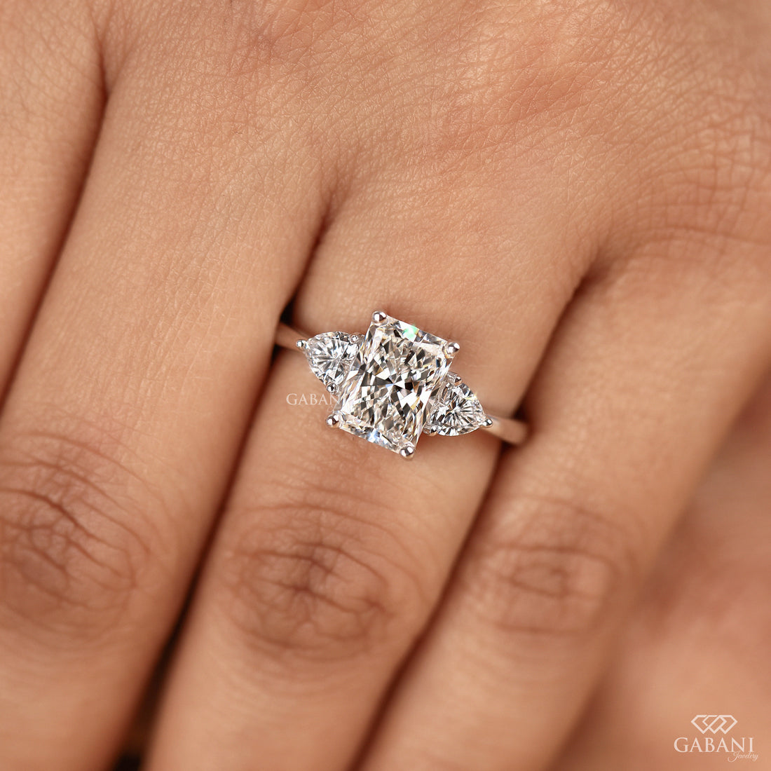 Radiant cut lab grown diamond three stone engagement ring with pear side diamonds, classic prong setting and polished gold band.