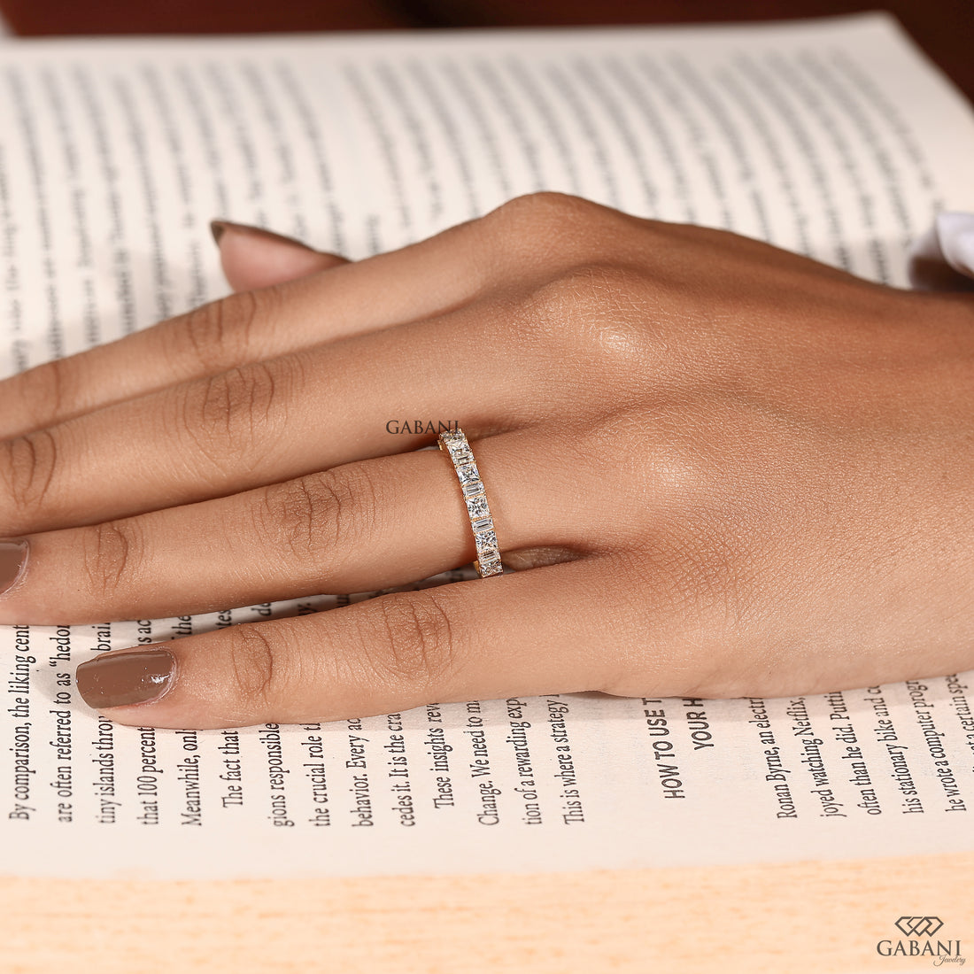 A close-up of a hand resting on an open book, wearing a slim gold band ring set with small clear stones. The brand name GABANI is subtly printed on the ring and appears again in the bottom right corner.