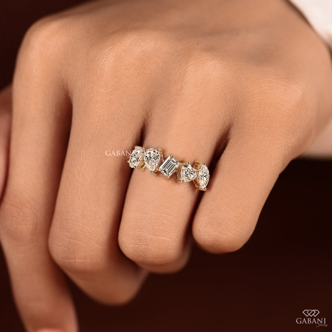A close-up of a hand wearing a delicate gold ring set with multiple clear gemstones, including a central emerald-cut stone. The brand name GABANI is visible as a watermark and in the bottom right corner.