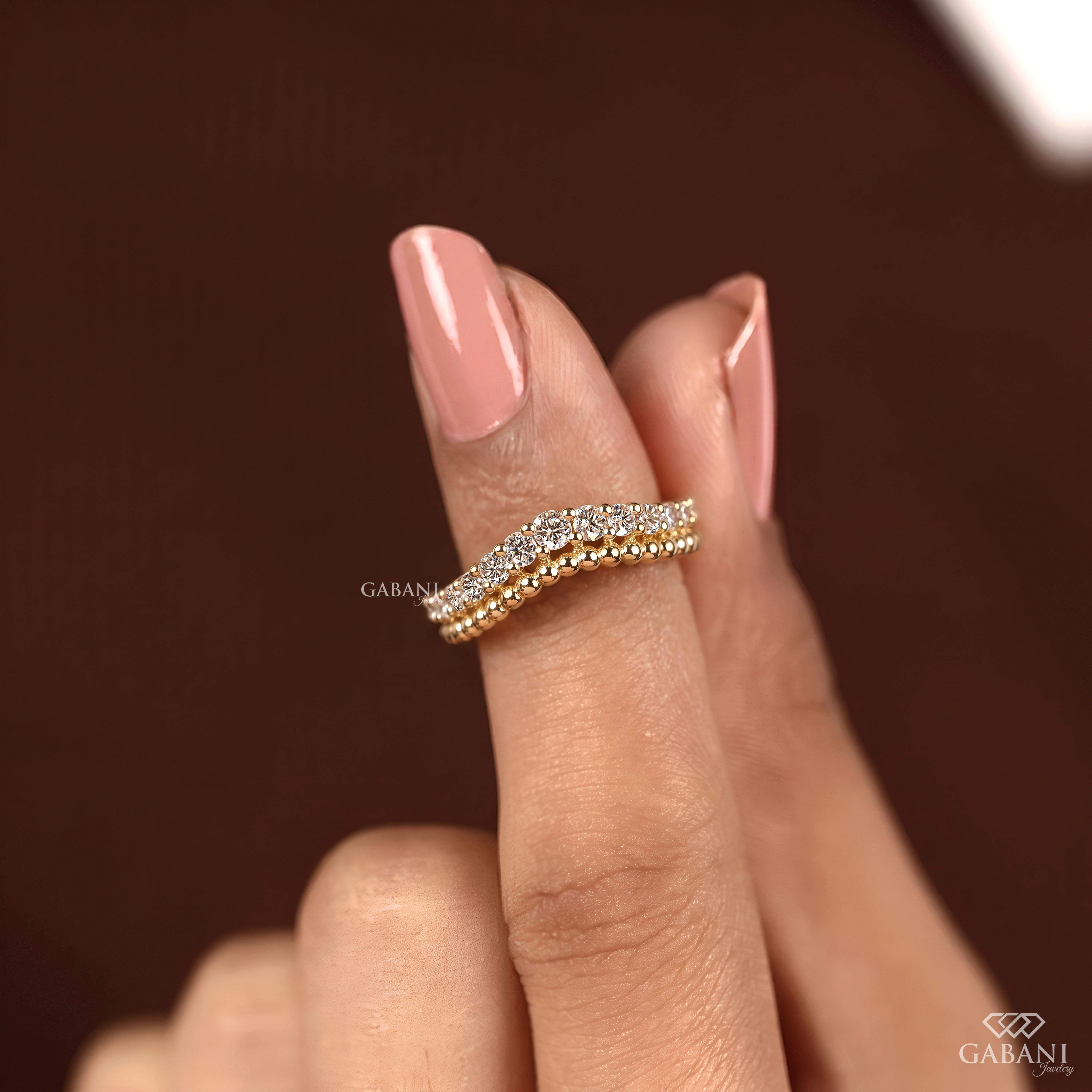 A close-up of a hand with pink manicured nails wearing a delicate wavy ring. The ring features a gold center channel flanked by rows of small clear stones in a silver-toned setting, set against a dark brown background.