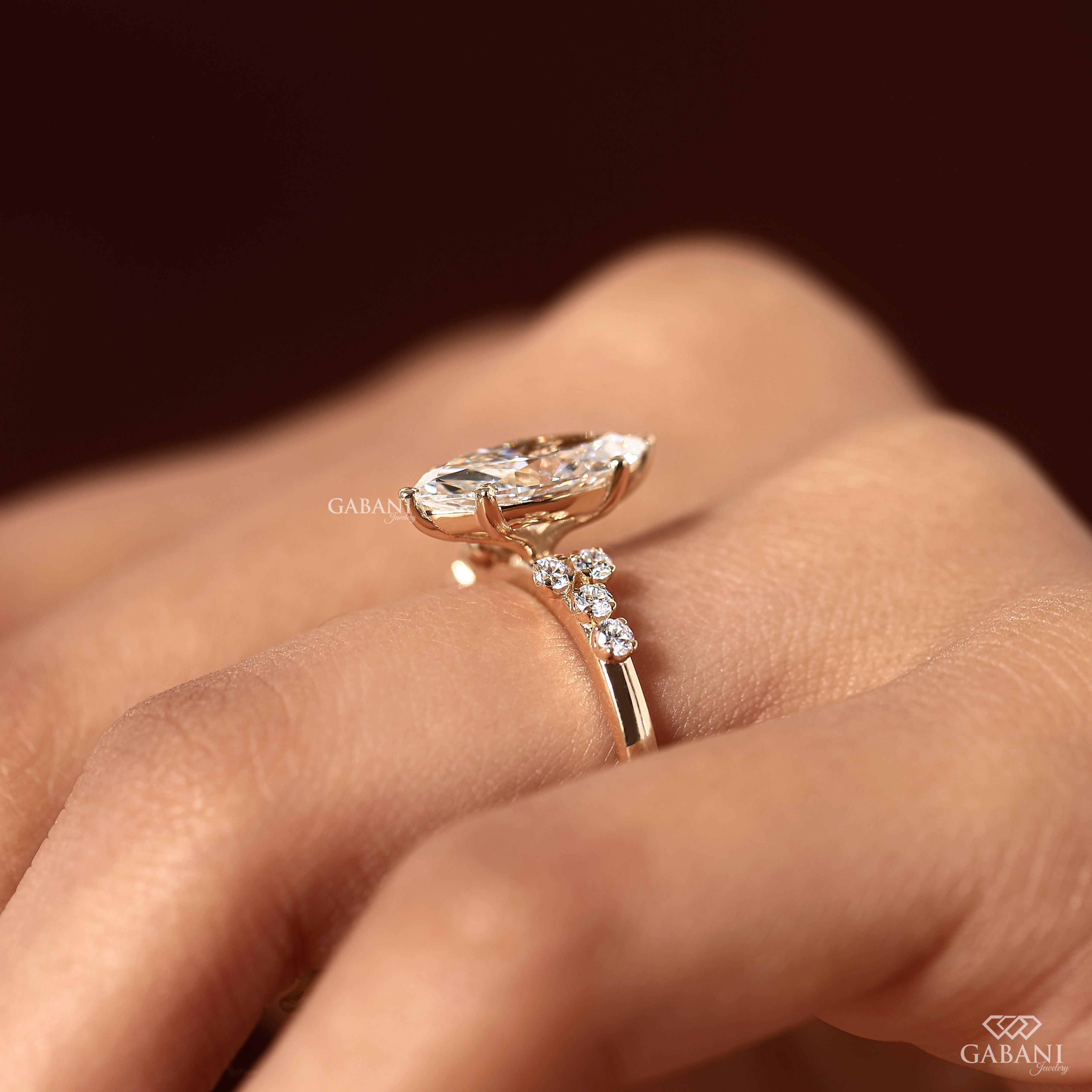 A close-up of a gold ring with a large marquise-cut center stone and smaller side stones, worn on a hand against a dark brown background. The brand name Gabani is visible as a watermark.