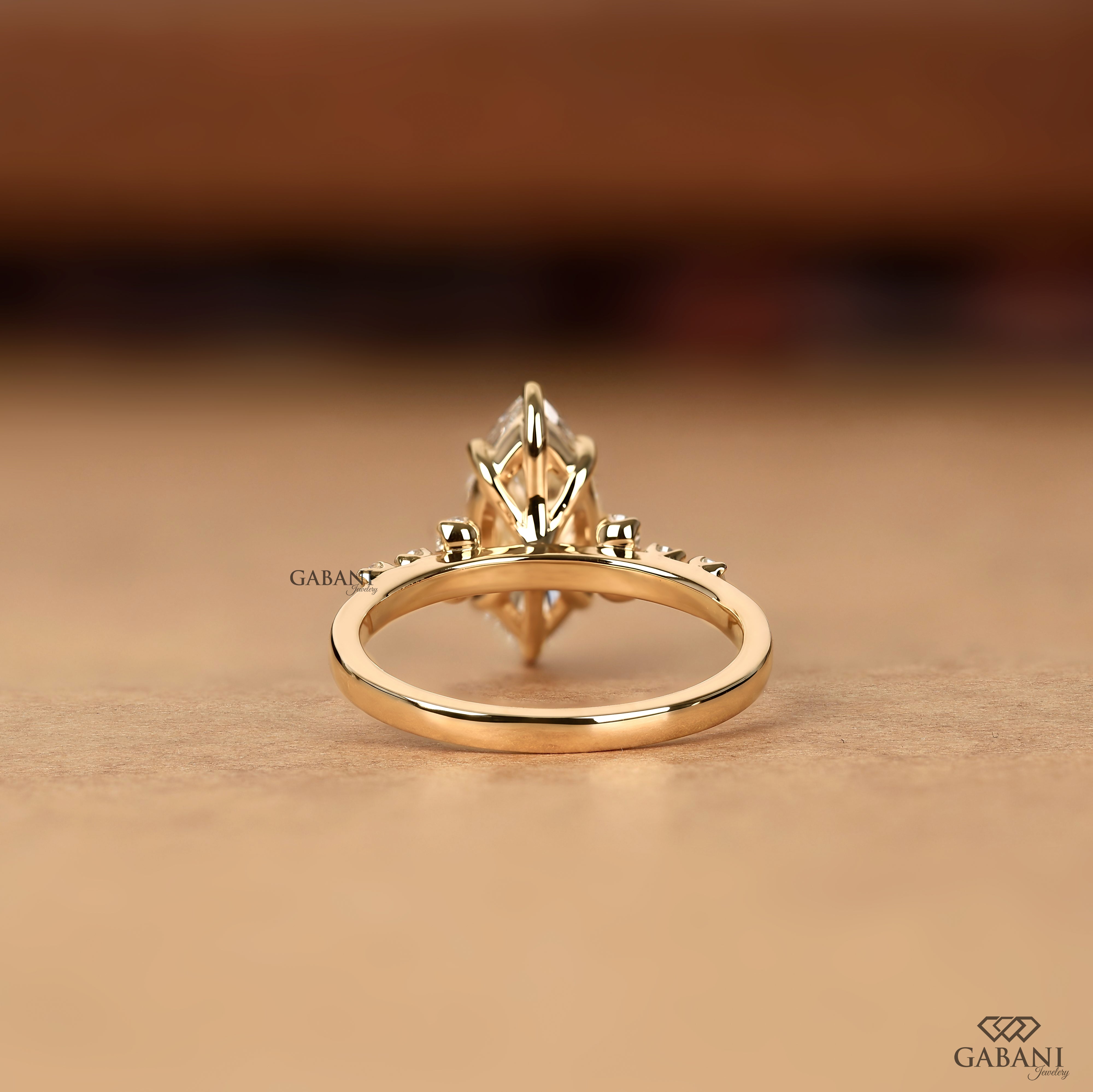 A gold ring with a marquise-cut center stone and small side stones, shown on a warm beige surface with a blurred brown background. The brand name 'GABANI' is visible as a watermark.