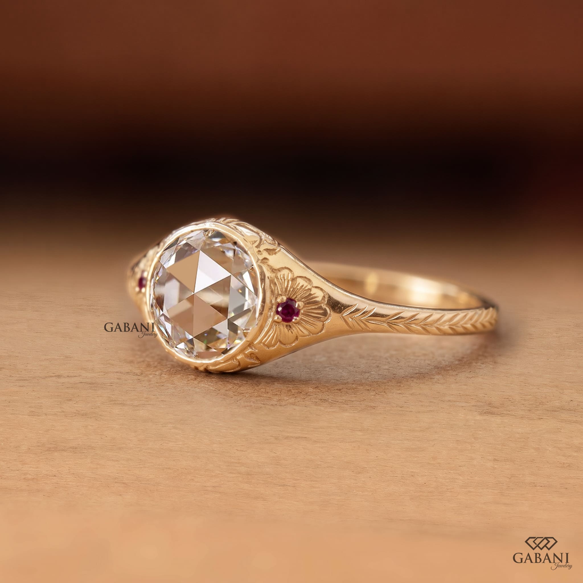 ALT Text: Round rose cut lab grown diamond engagement ring with ruby side accents, floral engraving, and bezel setting in 14K yellow gold.