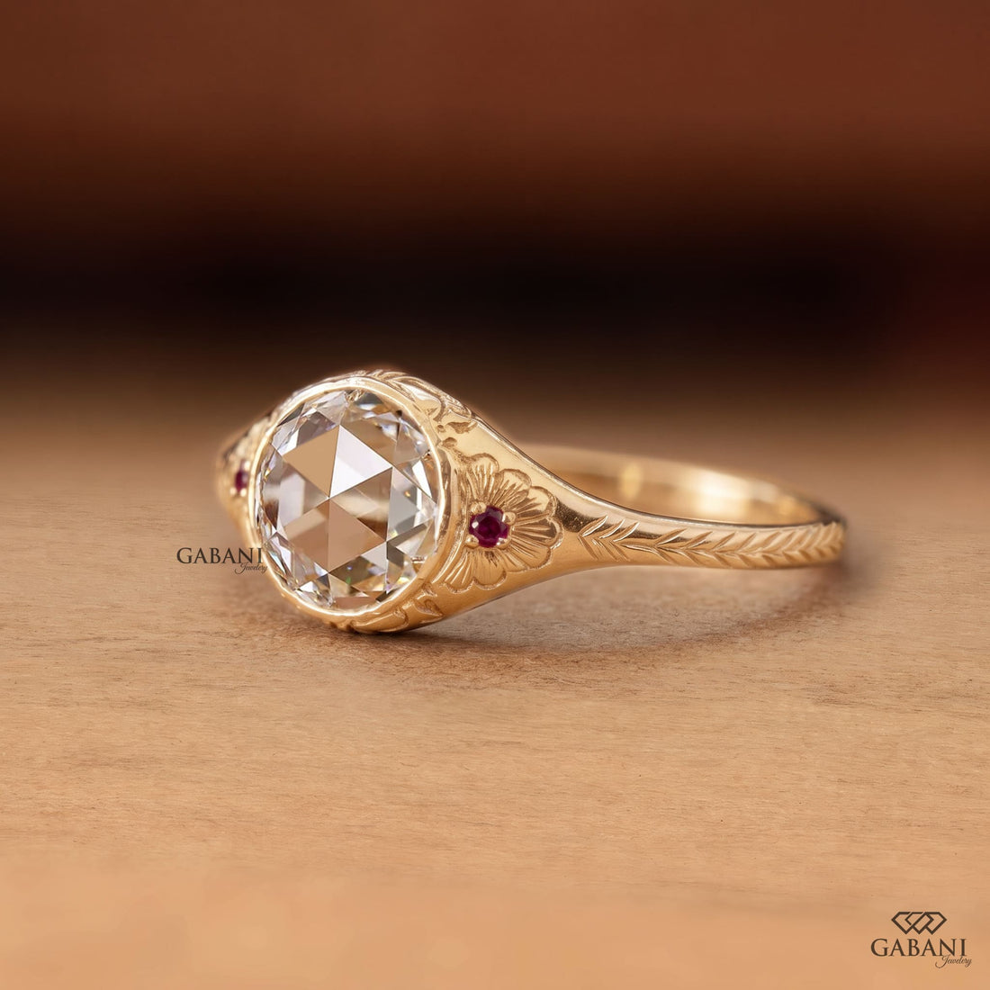 ALT Text: Round rose cut lab grown diamond engagement ring with ruby side accents, floral engraving, and bezel setting in 14K yellow gold.