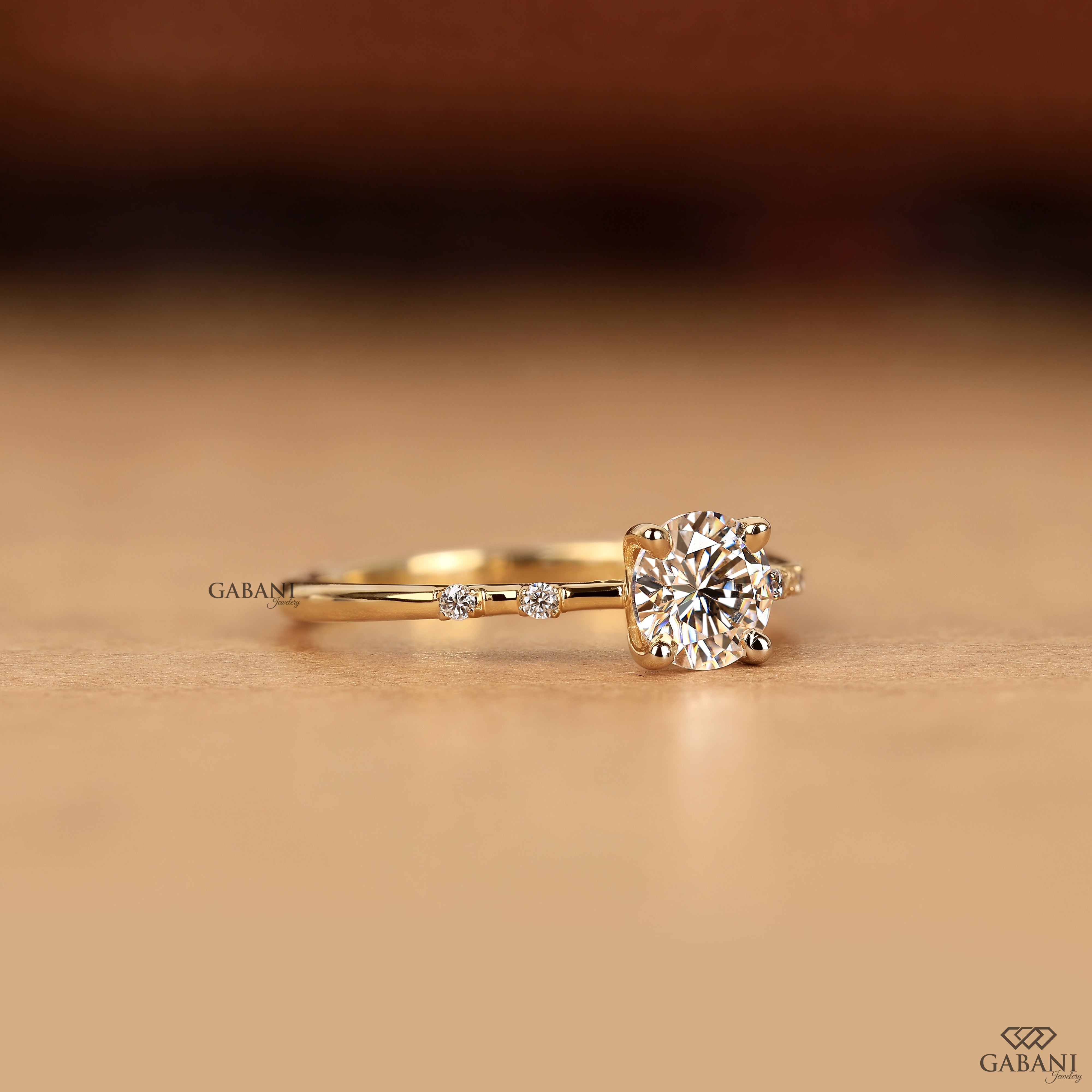 A yellow gold solitaire engagement ring with a round-cut diamond center stone and a small accent diamond on the band, resting on a warm beige surface.