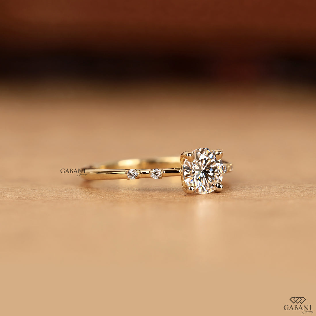 A yellow gold solitaire engagement ring with a round-cut diamond center stone and a small accent diamond on the band, resting on a warm beige surface.