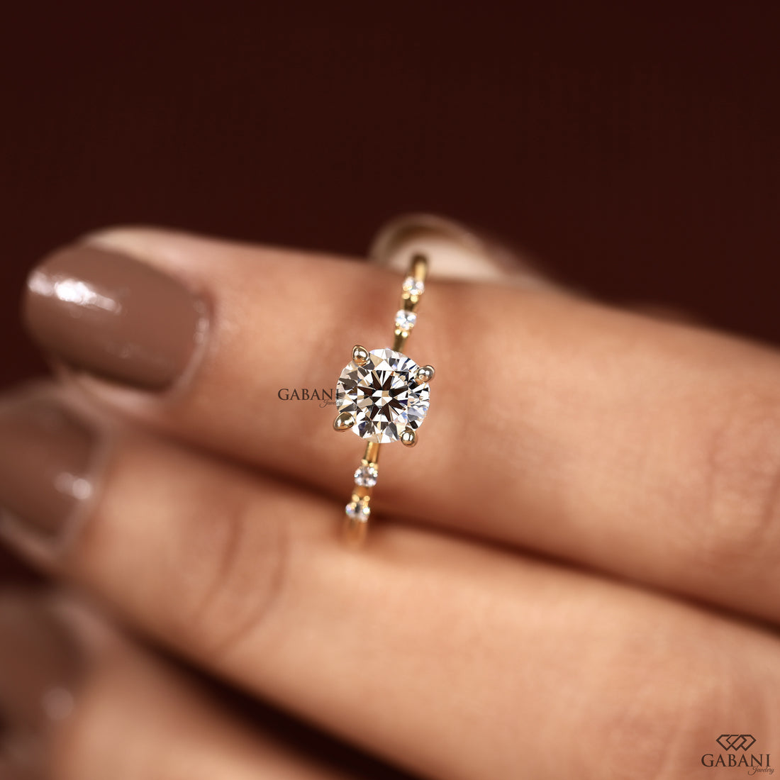 A gold ring with a large round-cut clear center stone and small accent stones, shown worn on a hand with brown manicured nails against a dark brown background.