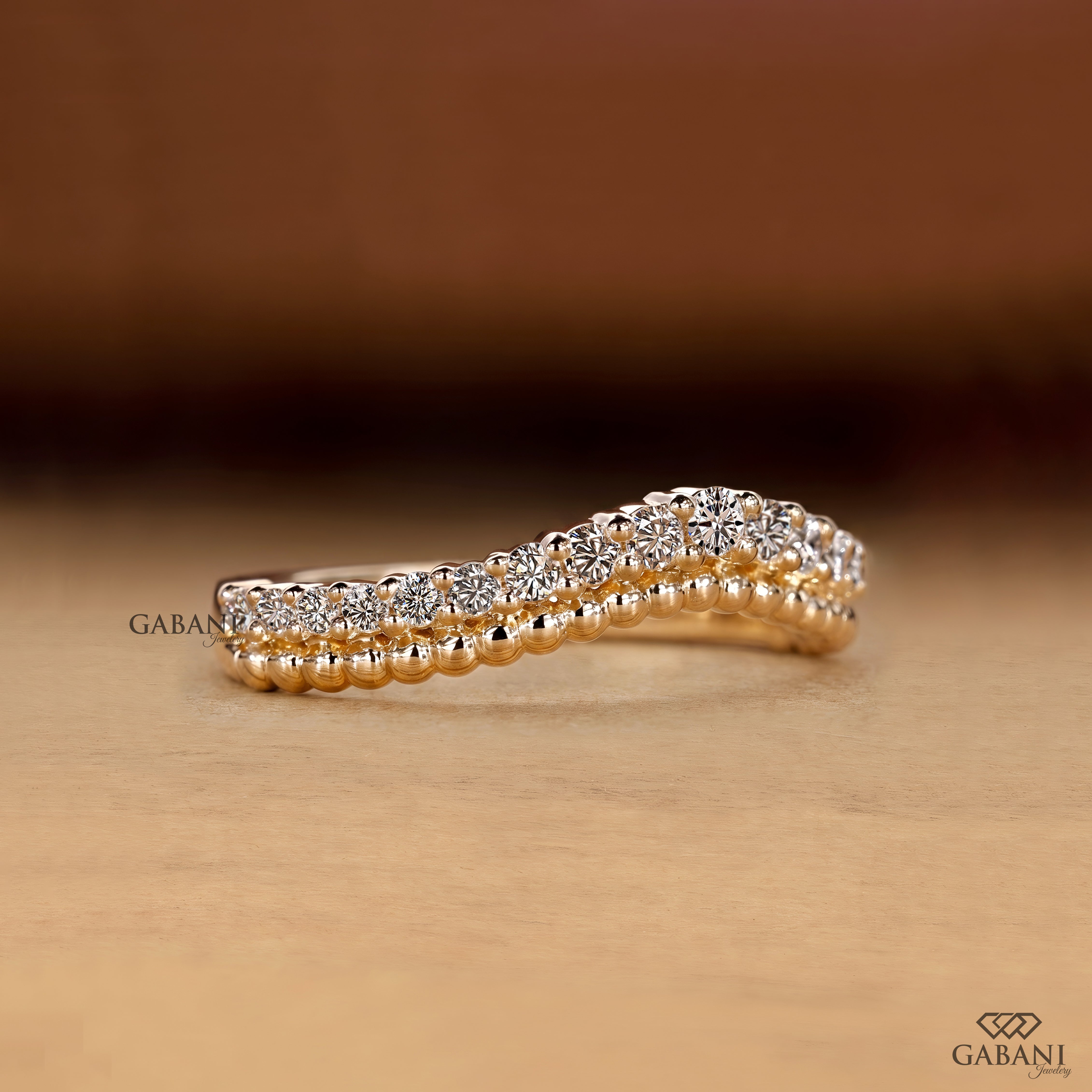 A close-up of a gold ring with a double row of sparkling clear gemstones, set against a warm brown and beige background. The brand name 'GABANI' is visible as a watermark and logo in the corners.