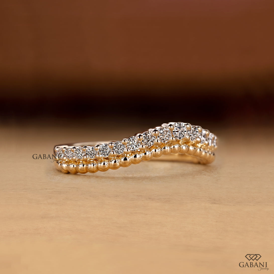 A close-up of a gold ring with a double row of sparkling clear gemstones, set against a warm brown and beige background. The brand name 'GABANI' is visible as a watermark and logo in the corners.