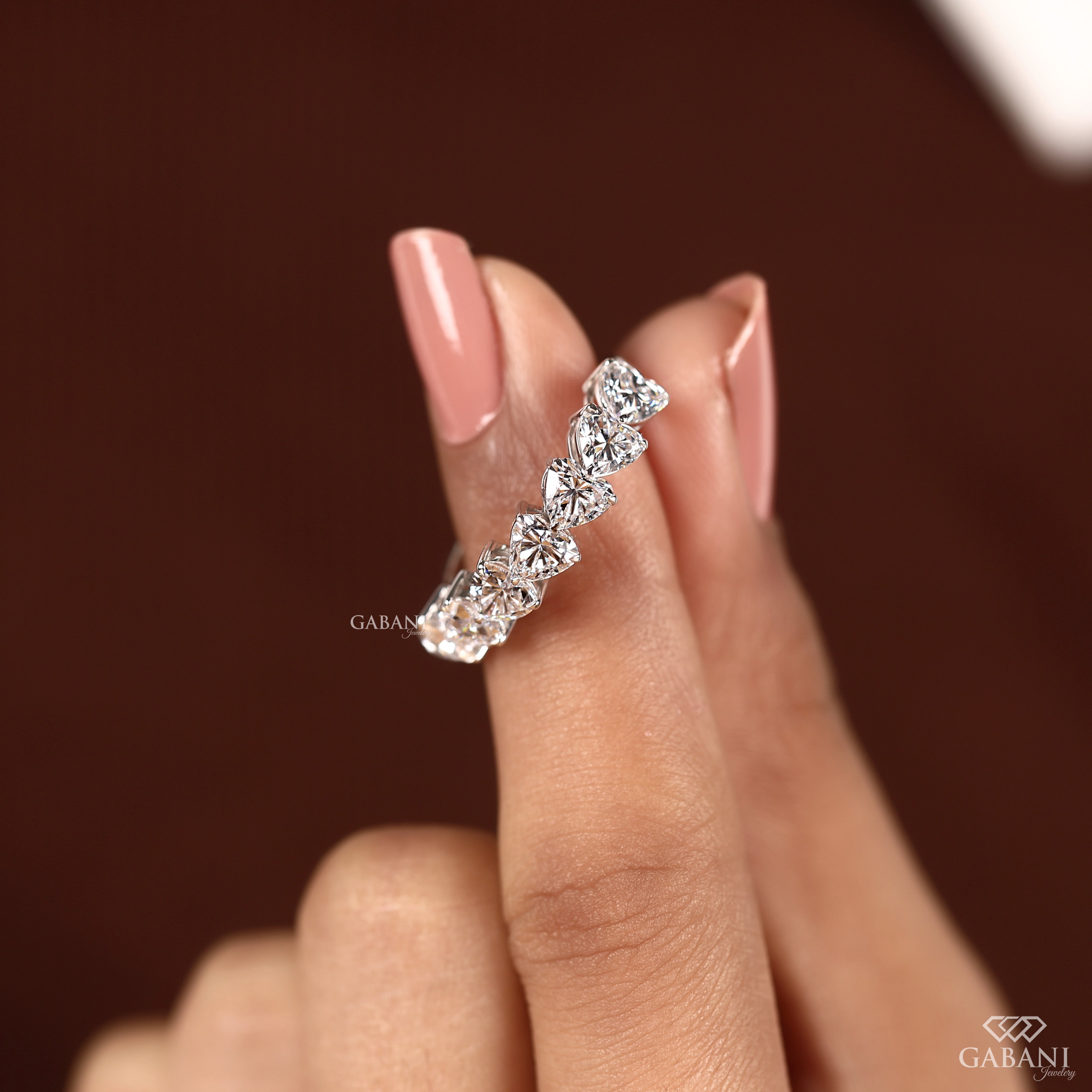 A close-up of a hand with pink manicured nails holding a silver-tone diamond band ring. The ring features a row of clear marquise-cut diamonds set in a silver band.