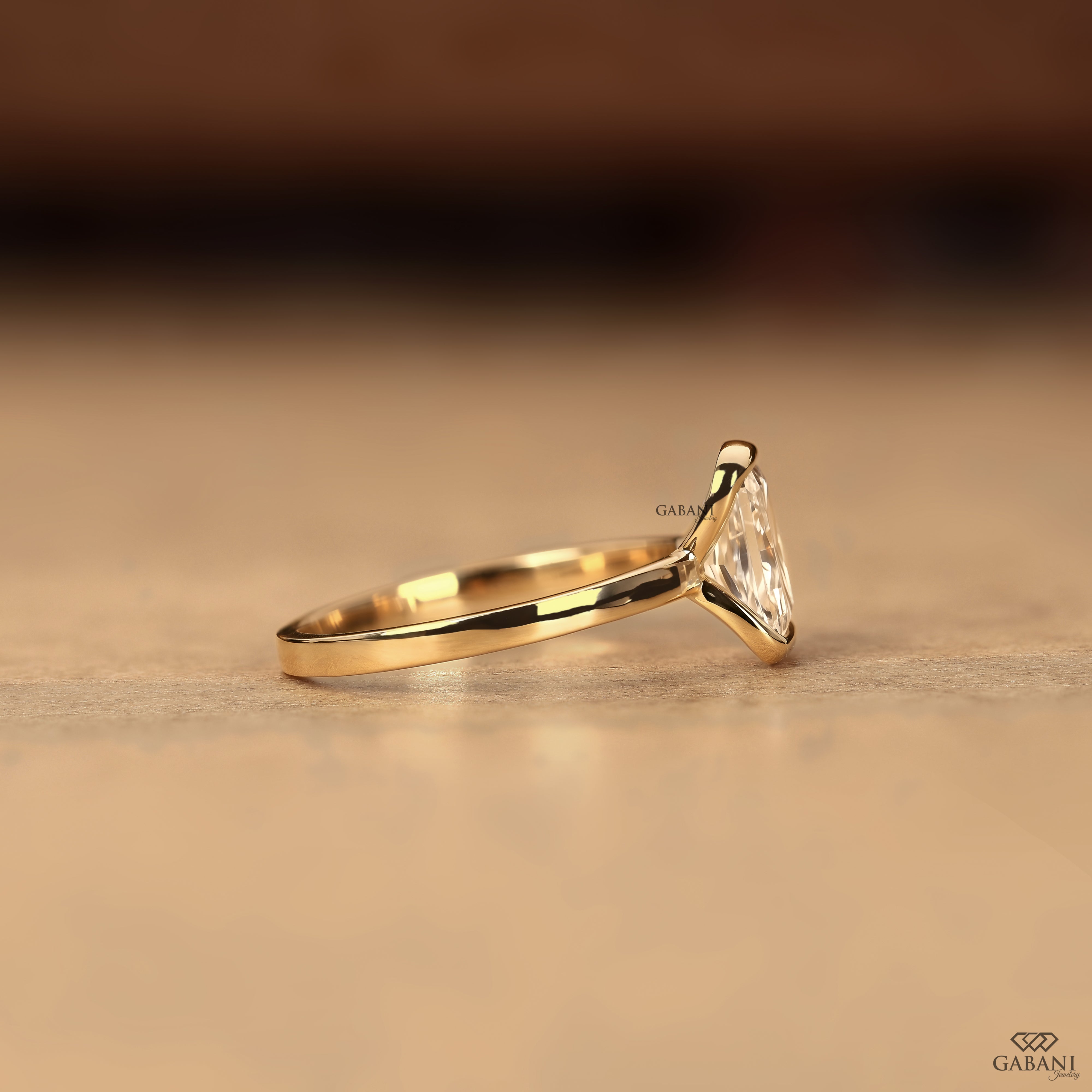 A gold ring with a triangular clear gemstone in a prong setting, resting on a textured beige surface with a blurred dark brown background.