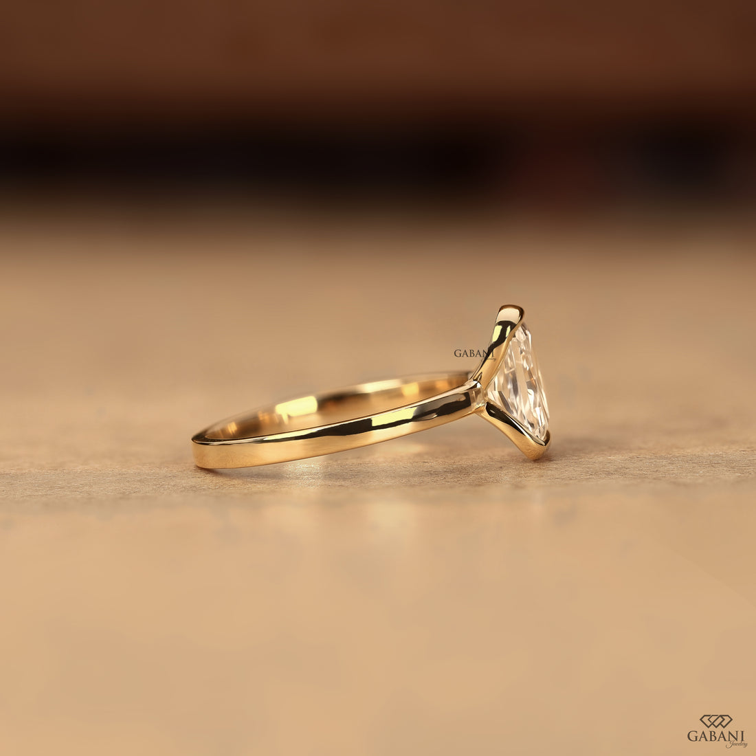 A gold ring with a triangular clear gemstone in a prong setting, resting on a textured beige surface with a blurred dark brown background.