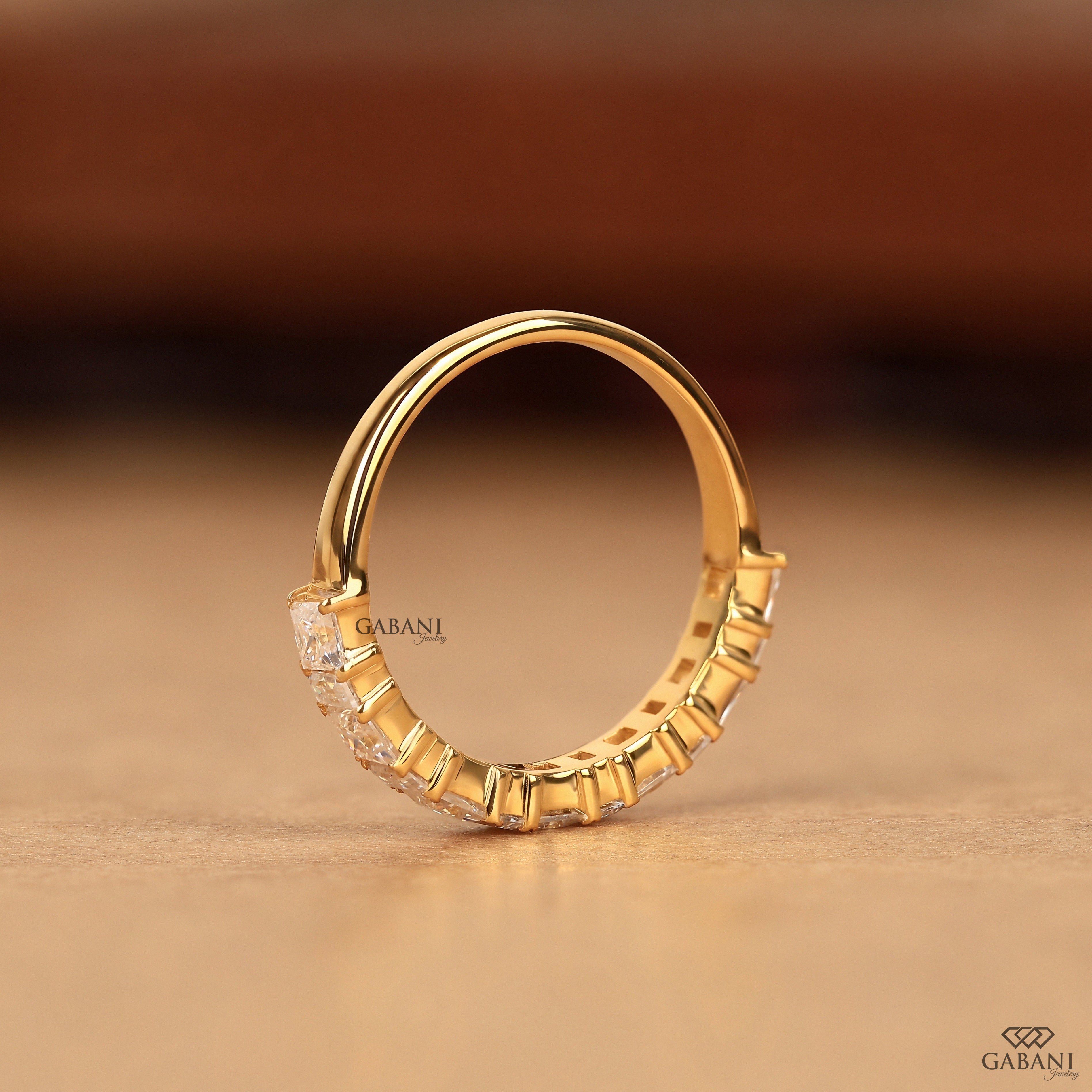 A gold hoop ring with a band featuring a row of small clear stones, photographed on a warm beige surface with a blurred brown background. The brand name GABANI is visible as a watermark.