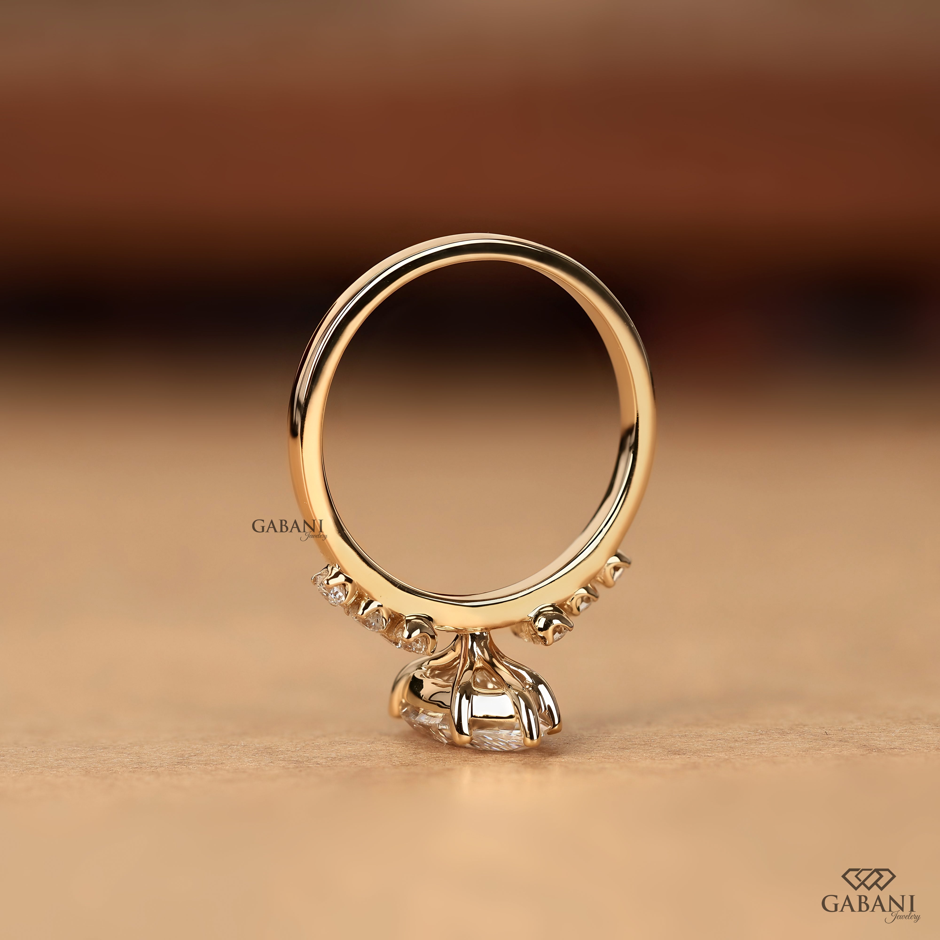 A gold ring with a large clear center stone and small accent stones along the band, photographed on a warm beige surface with a blurred brown background. The brand name GABANI appears as a watermark.
