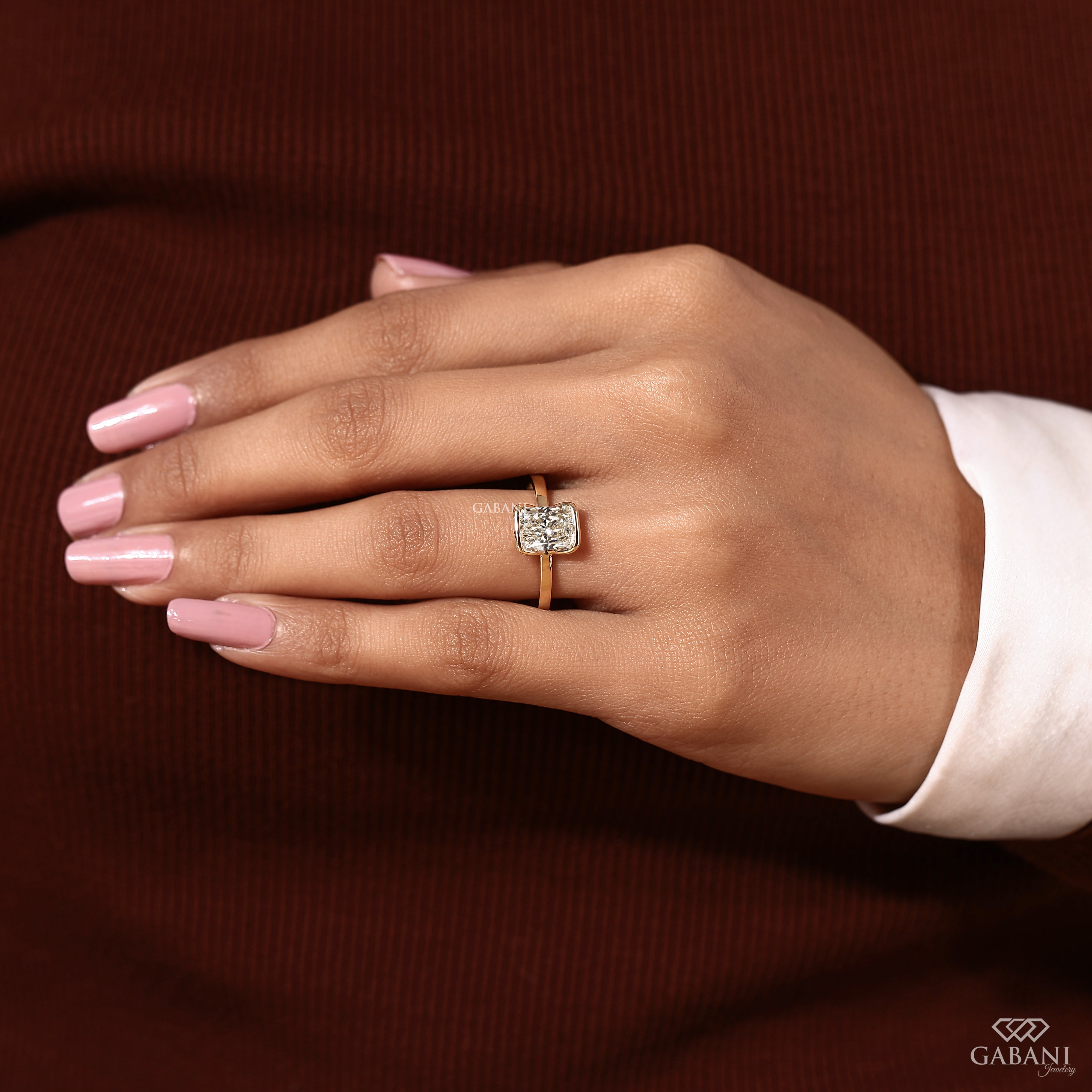 A close-up of a hand wearing a square-cut diamond ring on a gold band, set against a dark brown ribbed knit background and a white shirt cuff.