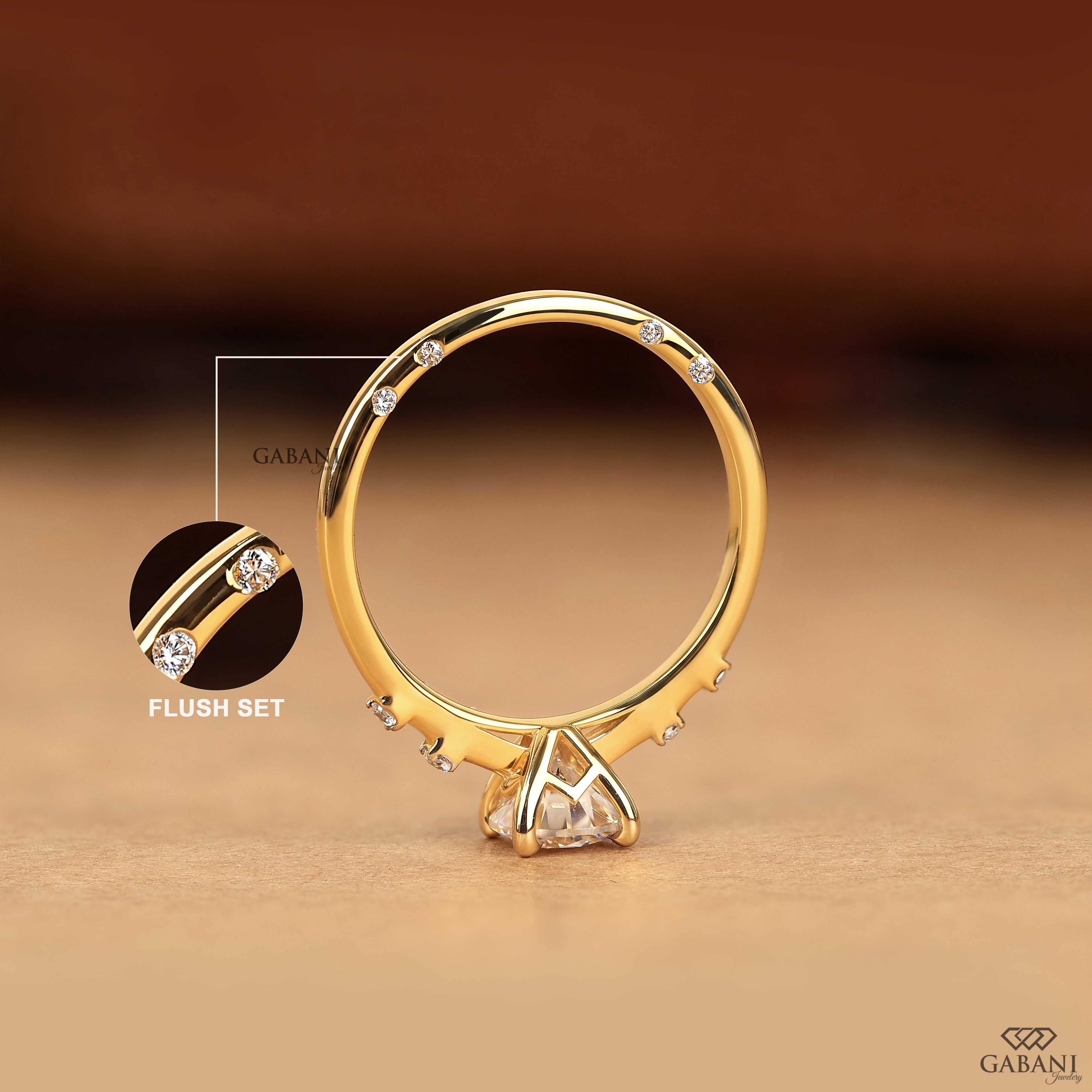 A gold ring with a triangular clear gemstone setting and small accent stones along the band, shown on a warm brown background. A circular inset highlights the flush-set detail, with the brand name GABANI visible.