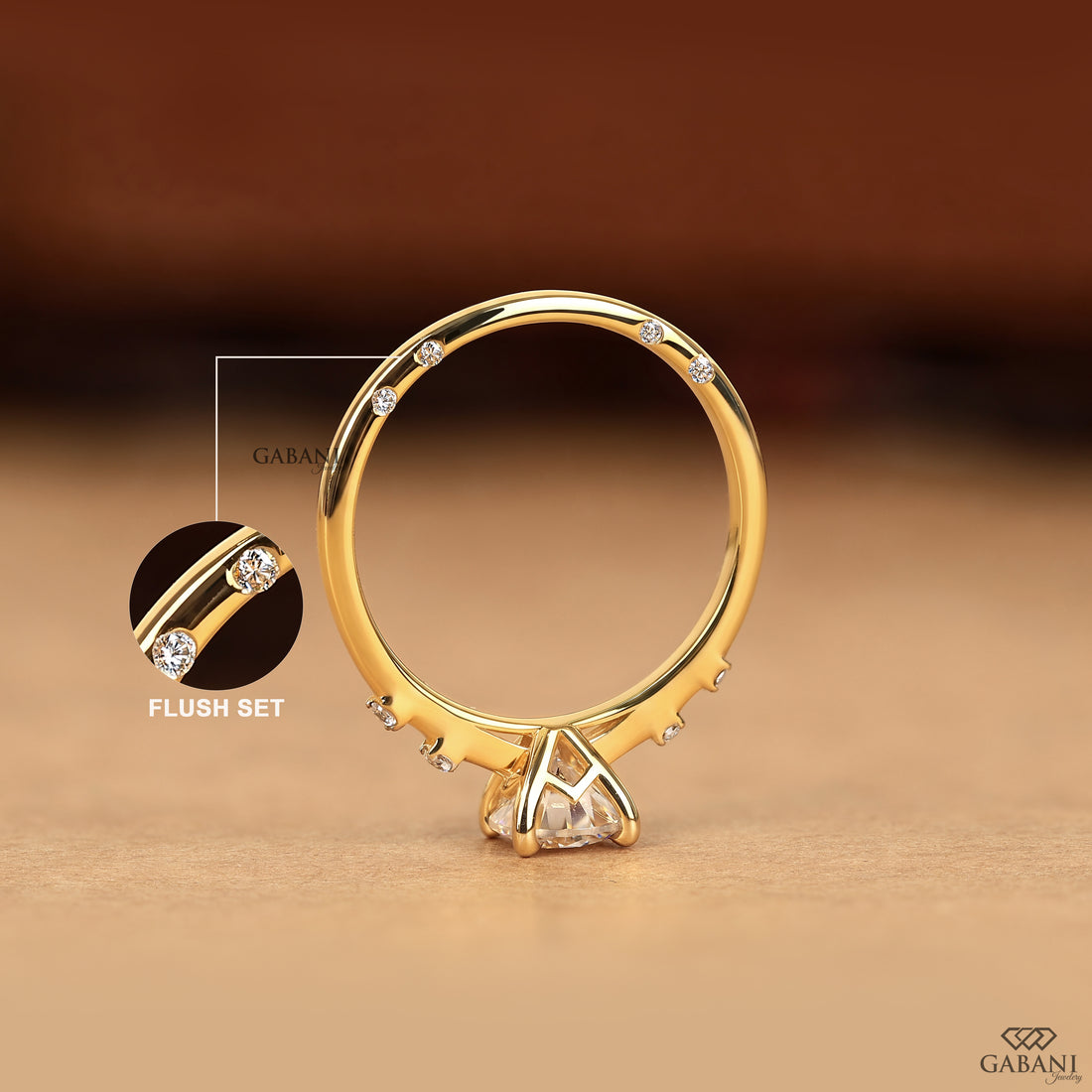 A gold ring with a triangular clear gemstone setting and small accent stones along the band, shown on a warm brown background. A circular inset highlights the flush-set detail, with the brand name GABANI visible.