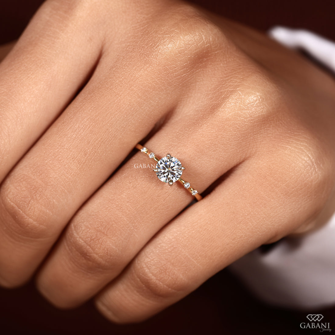 A close-up of a hand wearing a rose gold engagement ring with a round-cut diamond center stone and small side diamonds. The brand name GABANI is visible as a watermark and in the bottom right corner.