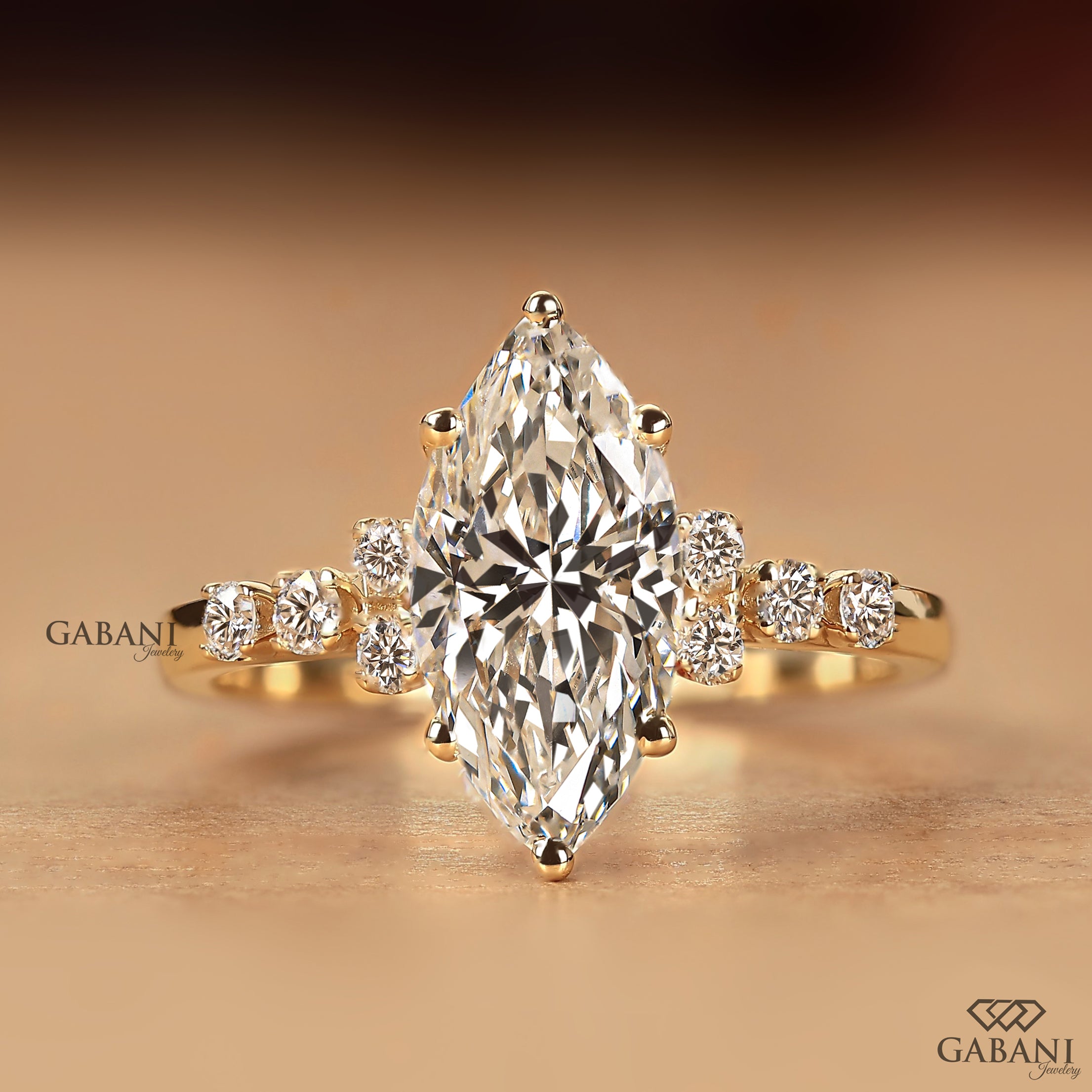 A yellow gold ring featuring a large marquise-cut diamond center stone with smaller round diamonds set along the shoulders, displayed on a warm beige background. The brand name 'GABANI' is visible as a watermark.