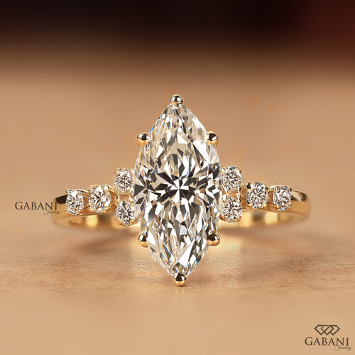 A yellow gold ring featuring a large marquise-cut diamond center stone with smaller round diamonds set along the shoulders, displayed on a warm beige background. The brand name 'GABANI' is visible as a watermark.