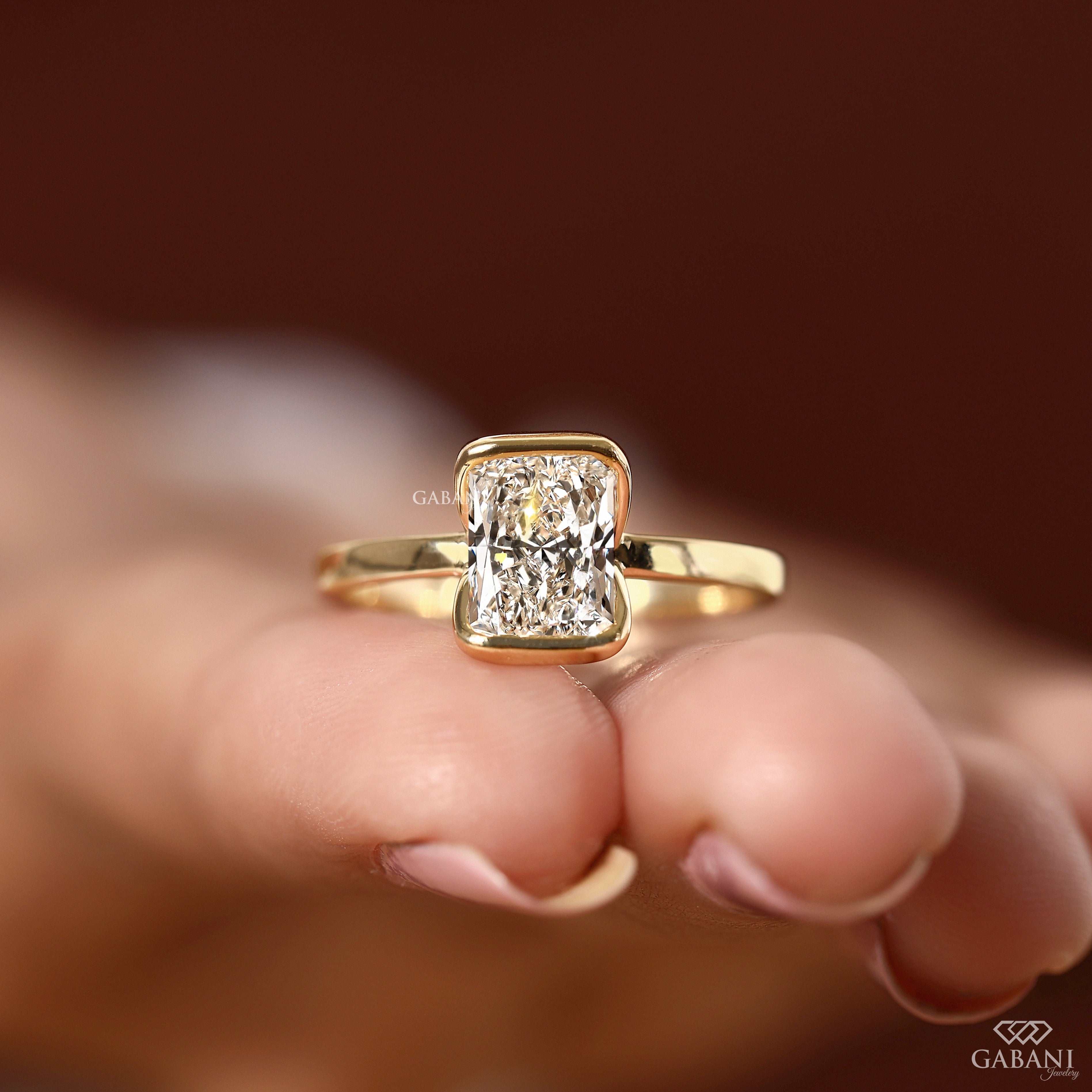 A yellow gold ring with a rectangular radiant-cut diamond set on a person's fingers, against a warm, blurred brown background. The brand name 'GABANI' is visible as a watermark and logo in the lower right corner.