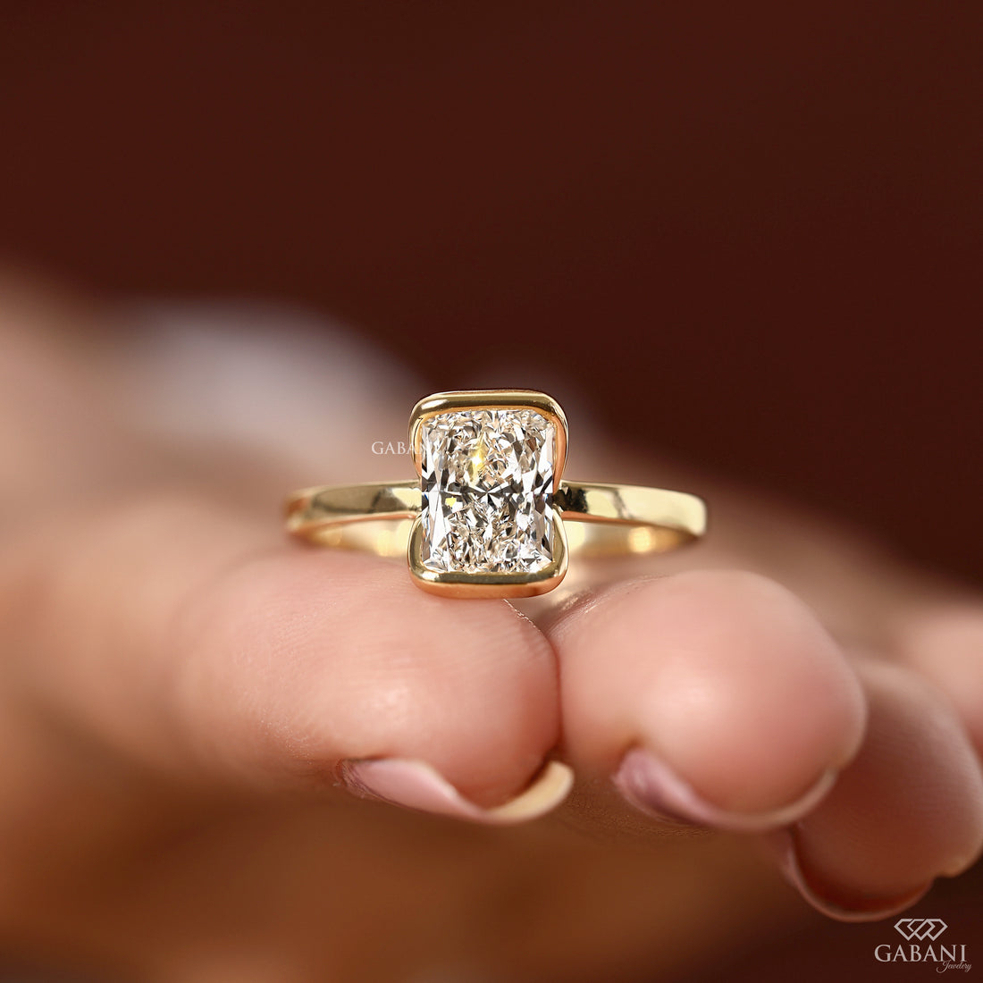 A yellow gold ring with a rectangular radiant-cut diamond set on a person's fingers, against a warm, blurred brown background. The brand name 'GABANI' is visible as a watermark and logo in the lower right corner.