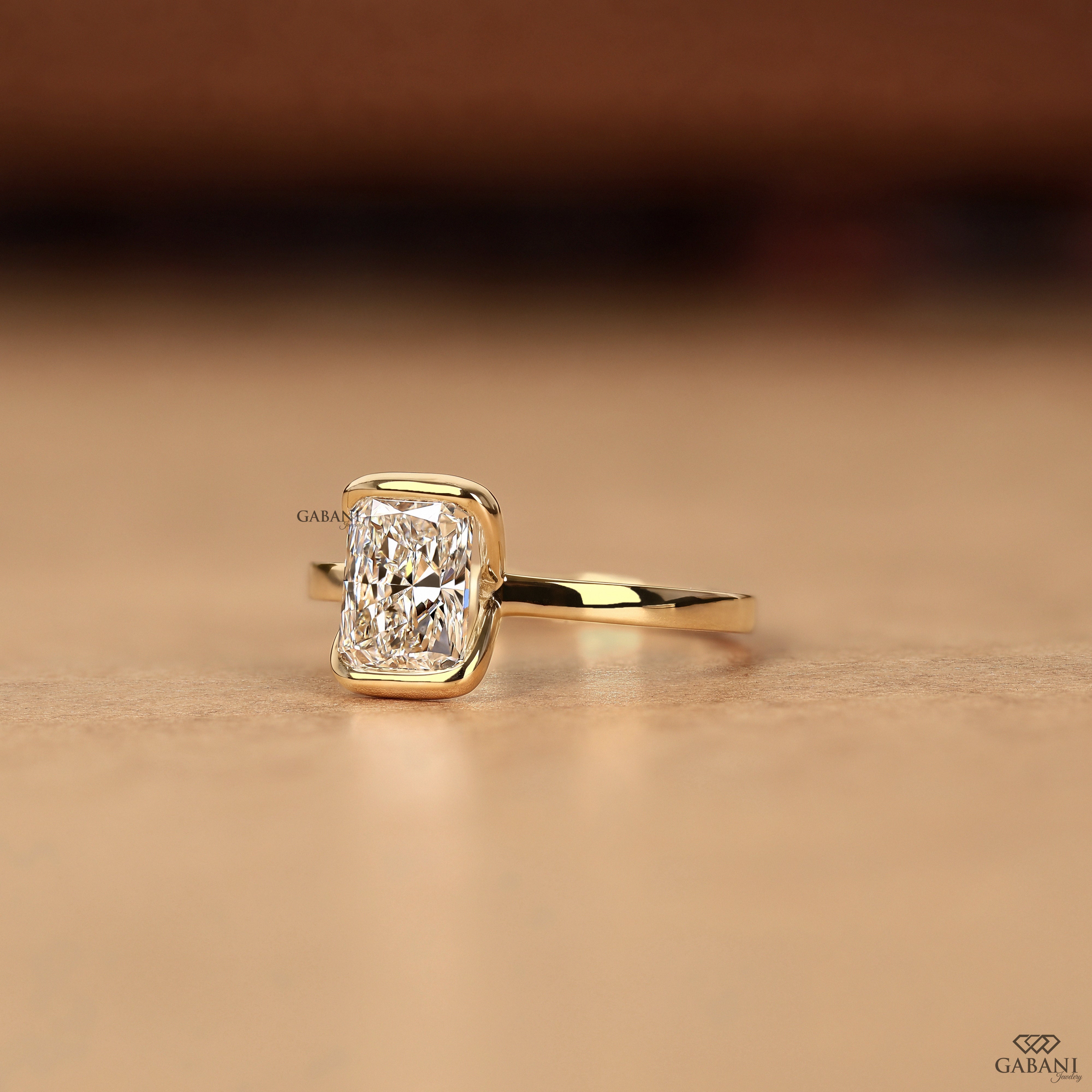 A yellow gold ring featuring a rectangular radiant-cut clear gemstone in a bezel setting, resting on a warm beige surface with a blurred brown background.