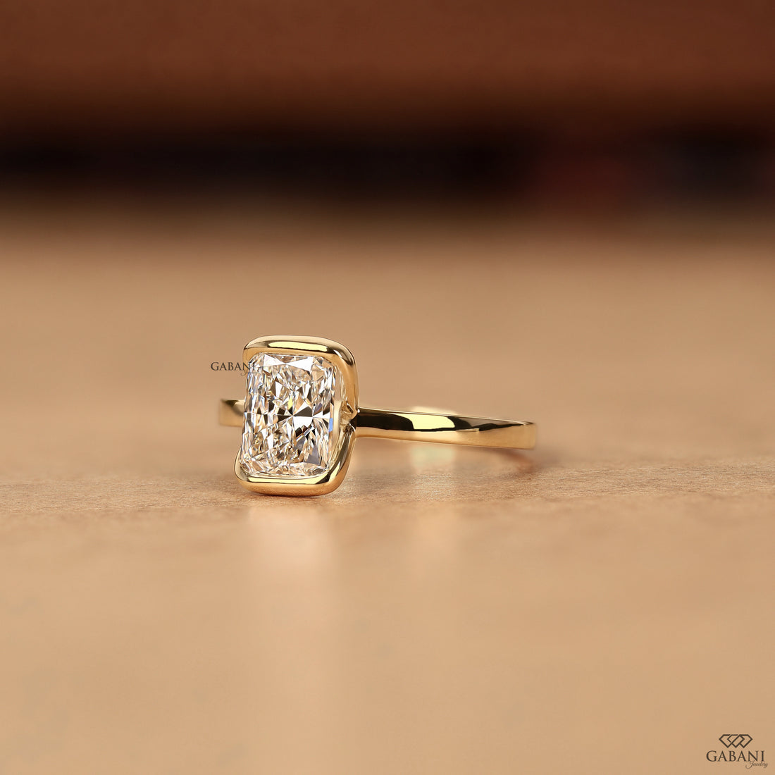 A yellow gold ring featuring a rectangular radiant-cut clear gemstone in a bezel setting, resting on a warm beige surface with a blurred brown background.