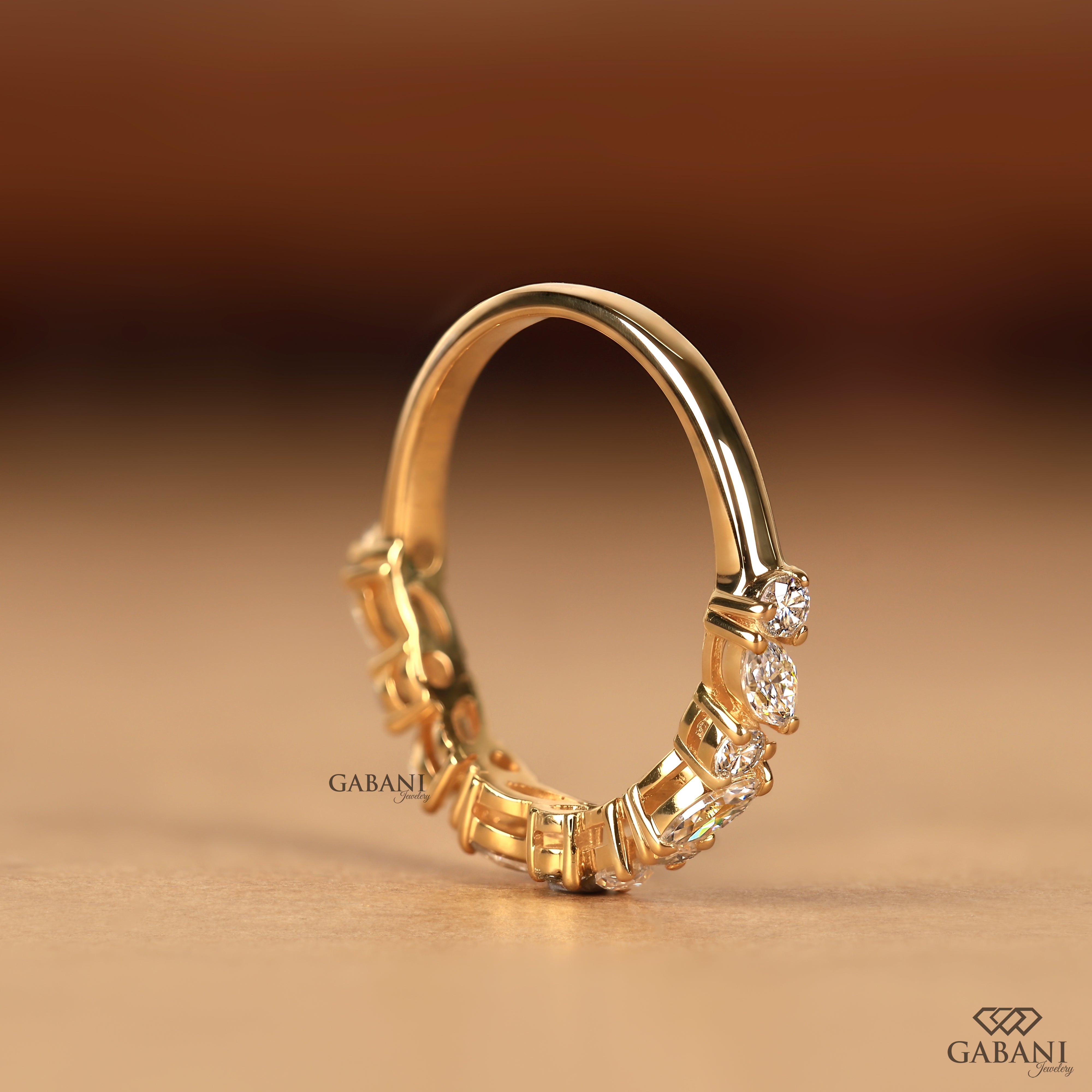 A gold ring with a curved band and a row of small clear stones, photographed on a warm beige surface with a blurred brown background. The brand name GABANI is visible near the base of the ring.