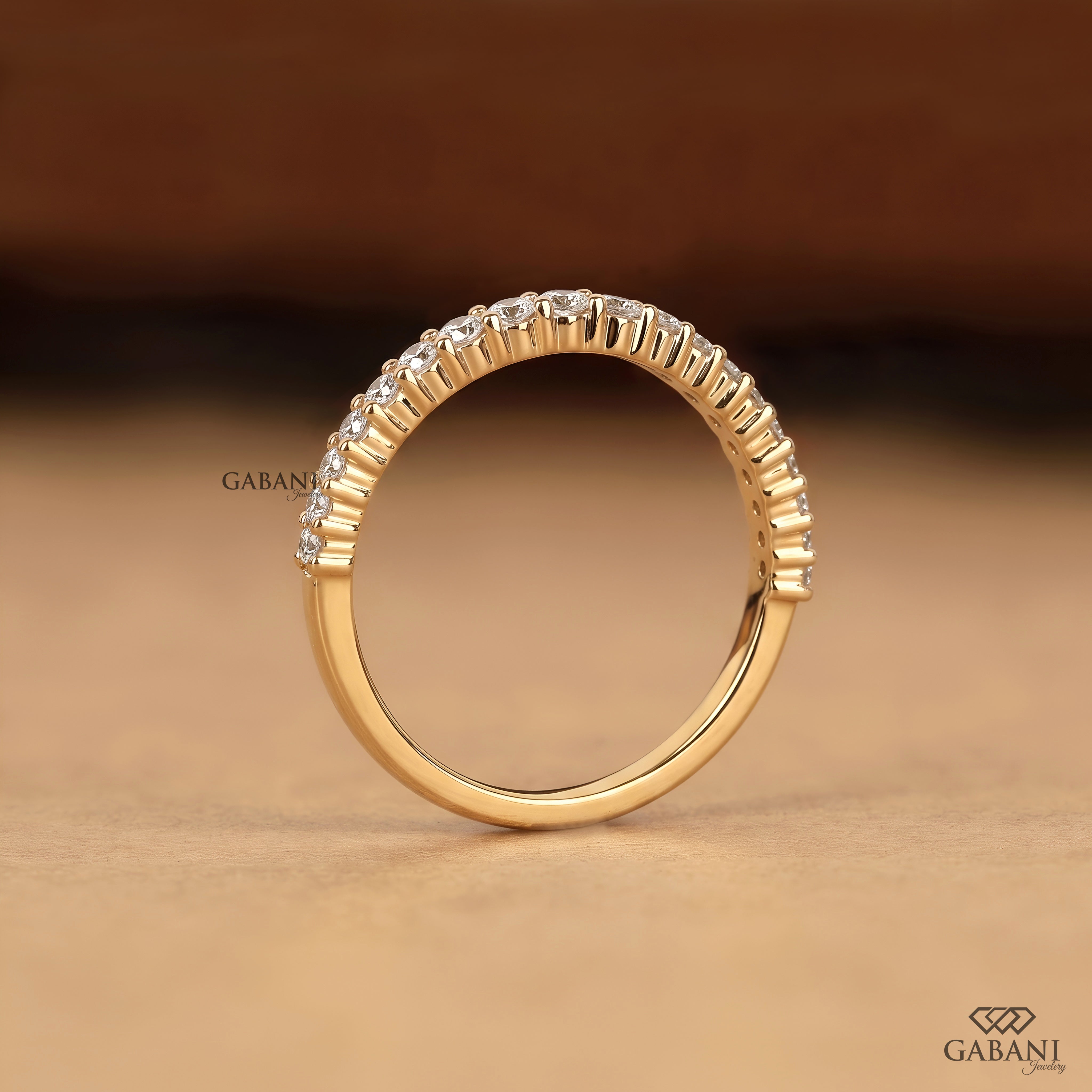 A gold ring with a curved row of small clear stones along the top half, photographed on a warm brown background. The brand name GABANI is visible as a watermark and logo in the corners.
