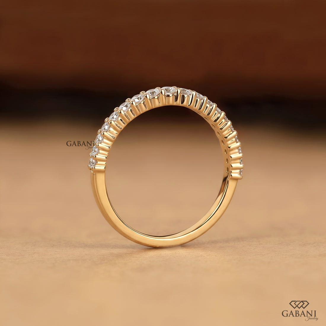 A gold ring with a curved row of small clear stones along the top half, photographed on a warm brown background. The brand name GABANI is visible as a watermark and logo in the corners.