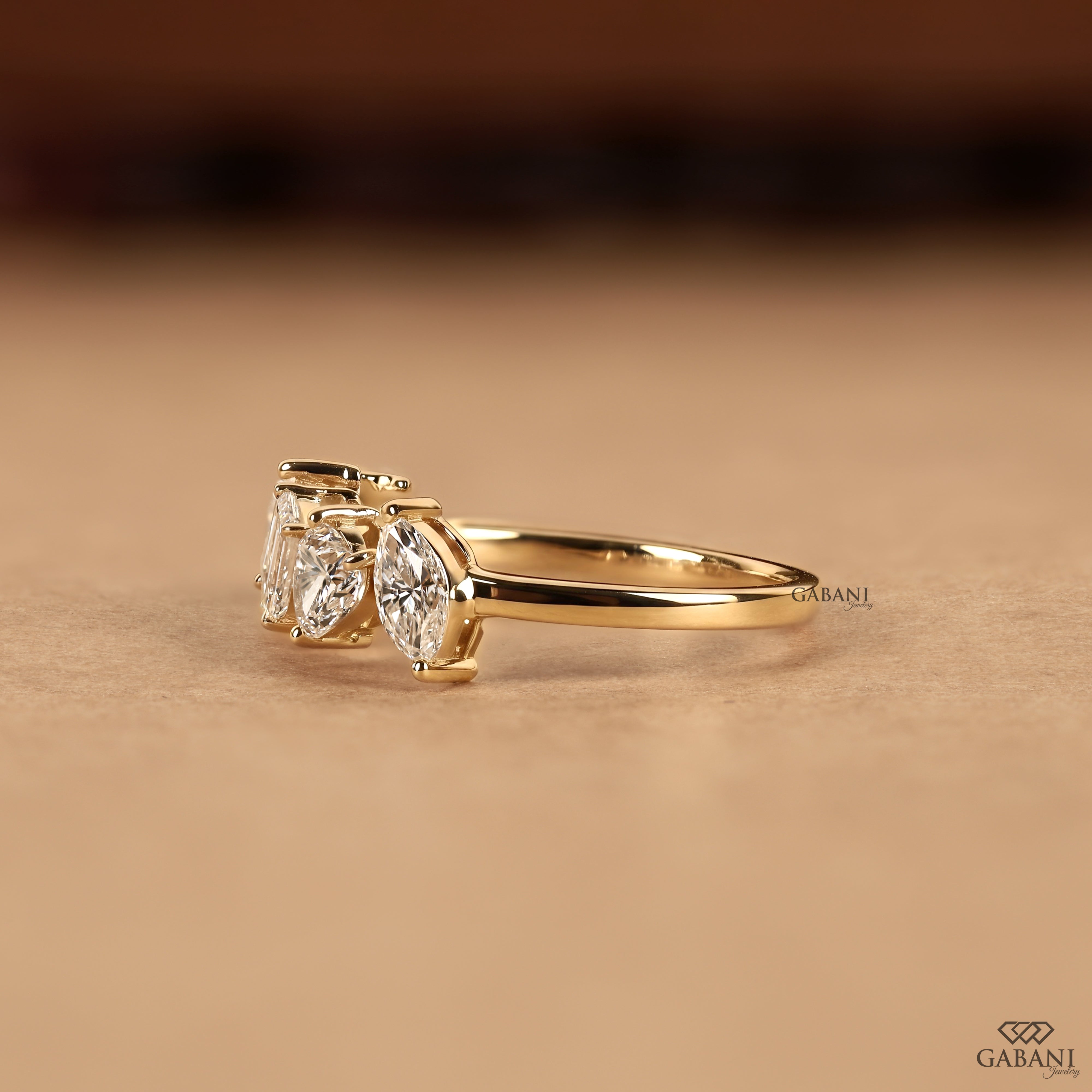 A gold ring with three marquise-cut clear gemstones, photographed on a warm beige surface with a blurred dark brown background. The brand name GABANI is visible near the ring and in the bottom right corner.