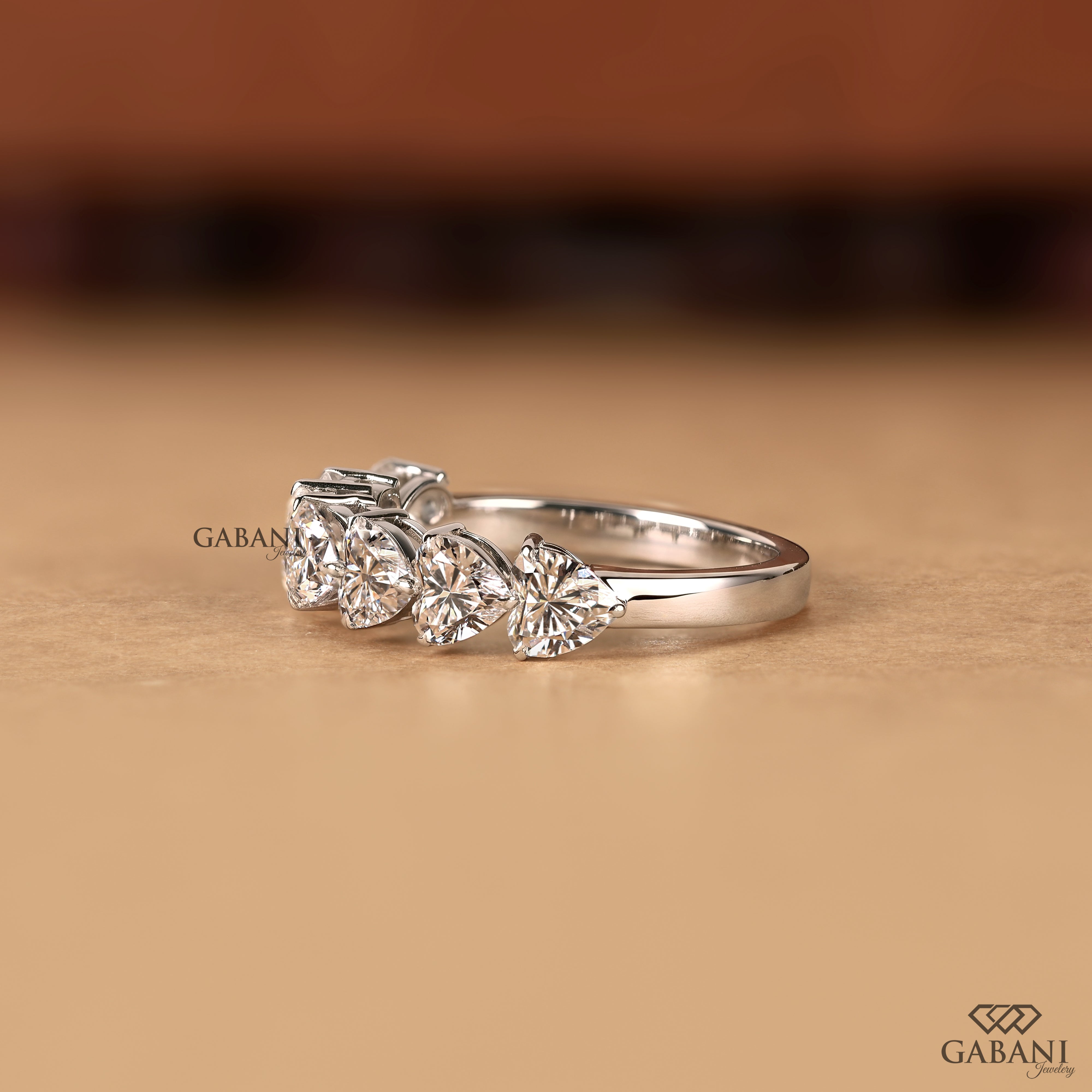 A silver ring with three clear heart-shaped gemstones, photographed on a warm beige surface with a blurred brown background. The brand name GABANI is visible as a watermark.